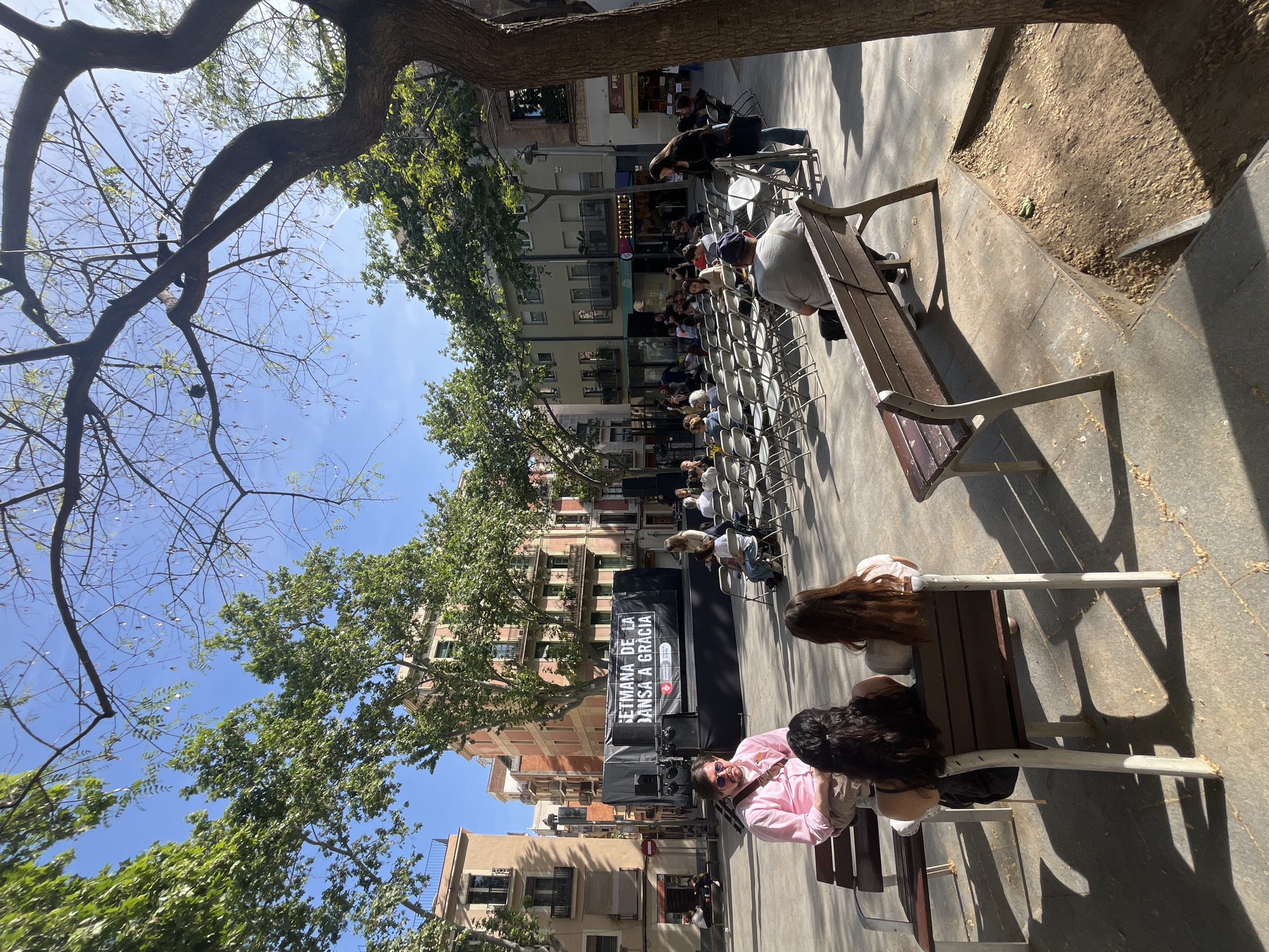 An outdoor public square, likely in the Gràcia district of Barcelona, Spain, is depicted during a sunny event. The scene shows numerous people gathered, mostly seated in rows of white foldable chairs facing a central stage. In the immediate foreground, individuals are seated on wooden and metal benches, observing the activity.
A prominent black stage is set up at the far end of the seating area, flanked by large speakers. A white banner across the stage displays the text "SETMANA DE LA DANSA A GRÀCIA" (Dance Week in Gràcia), indicating a dance-related festival or event. Below this, smaller text reads "GRÀCIA ES MOU" (Gràcia Moves) along with several organizational or sponsor logos.
The day is clear and bright, with a vivid blue sky overhead. Strong sunlight casts distinct shadows across the paved ground, suggesting it is mid-day to early afternoon. Large trees with both verdant green foliage and some bare branches frame the square, providing dappled shade. Multi-story buildings with balconies, characteristic of European urban architecture, line the perimeter of the plaza. The atmosphere appears to be calm, with attendees focused on the stage.