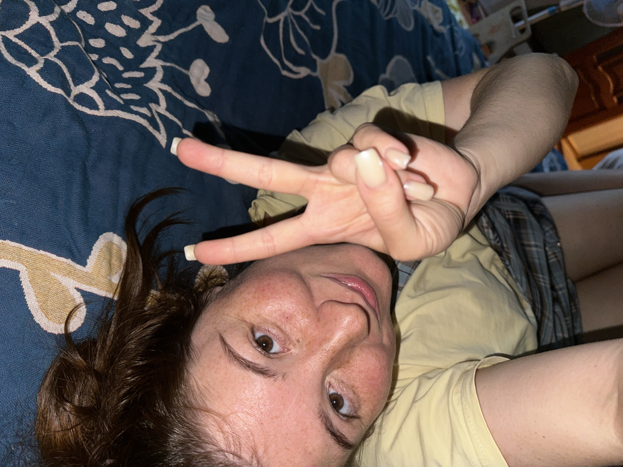 The image shows a young woman lying on a bed with a patterned blue and white comforter. She is wearing a yellow t-shirt and plaid shorts. Her face is partially visible, and she is making a peace sign with her right hand. The lighting suggests an indoor environment. There is no information in the image to determine its location in Nizhny Novgorod, Russia.