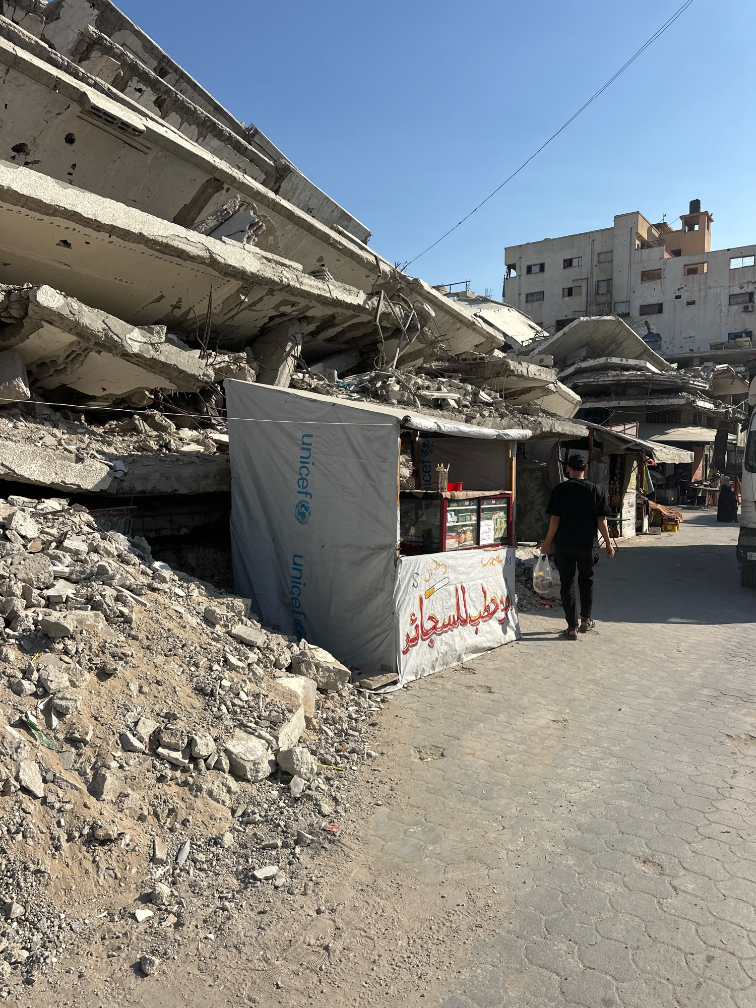 In the city of Gaza, Palestinian Territories, a man in a black t-shirt and black pants walks past a UNICEF-branded stall positioned next to a pile of rubble from a destroyed building. The stall has a tarp covering its front and displays various products inside. A sign with Arabic script is visible on the stall's front. Behind the stall, remnants of a collapsed concrete structure loom large against a clear blue sky. In the background, other buildings, some appearing damaged, are visible. The ground is paved with interlocking stones, showing signs of wear and some small indentations. Debris from the destroyed building is scattered on the left side of the image.