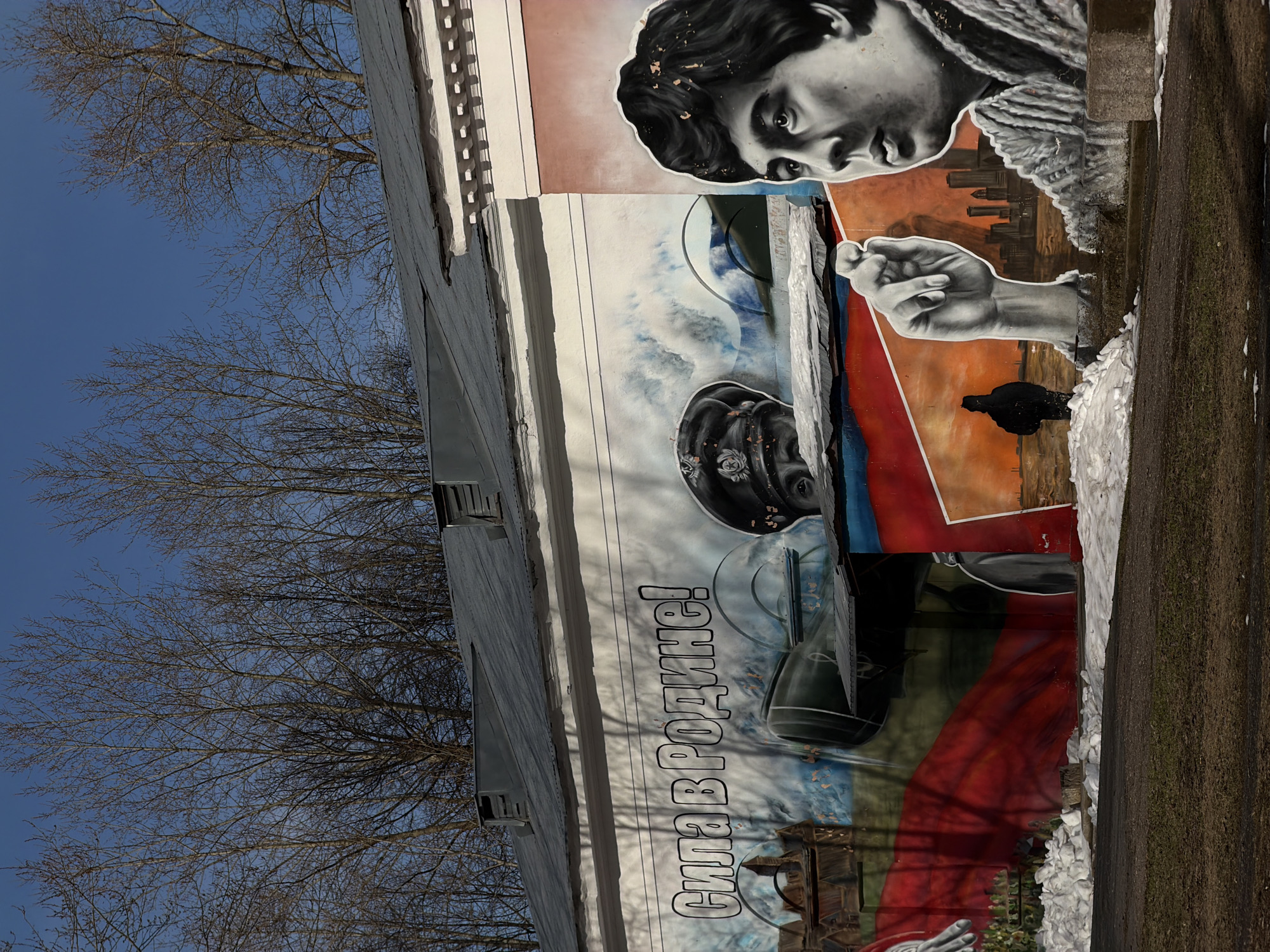 A mural on a building in Perm, Russia, features a black and white portrait of a young person looking down. The mural also includes a white hand gesturing, a cityscape silhouette in orange and brown tones, and a red, white, and blue section containing what appears to be a photograph of military equipment. The Russian text "Сила в Родине!" (Strength is in the Motherland!) is written vertically on the building. The building is located near trees without leaves under a blue sky. A snow-covered sidewalk runs along the right side of the building.
