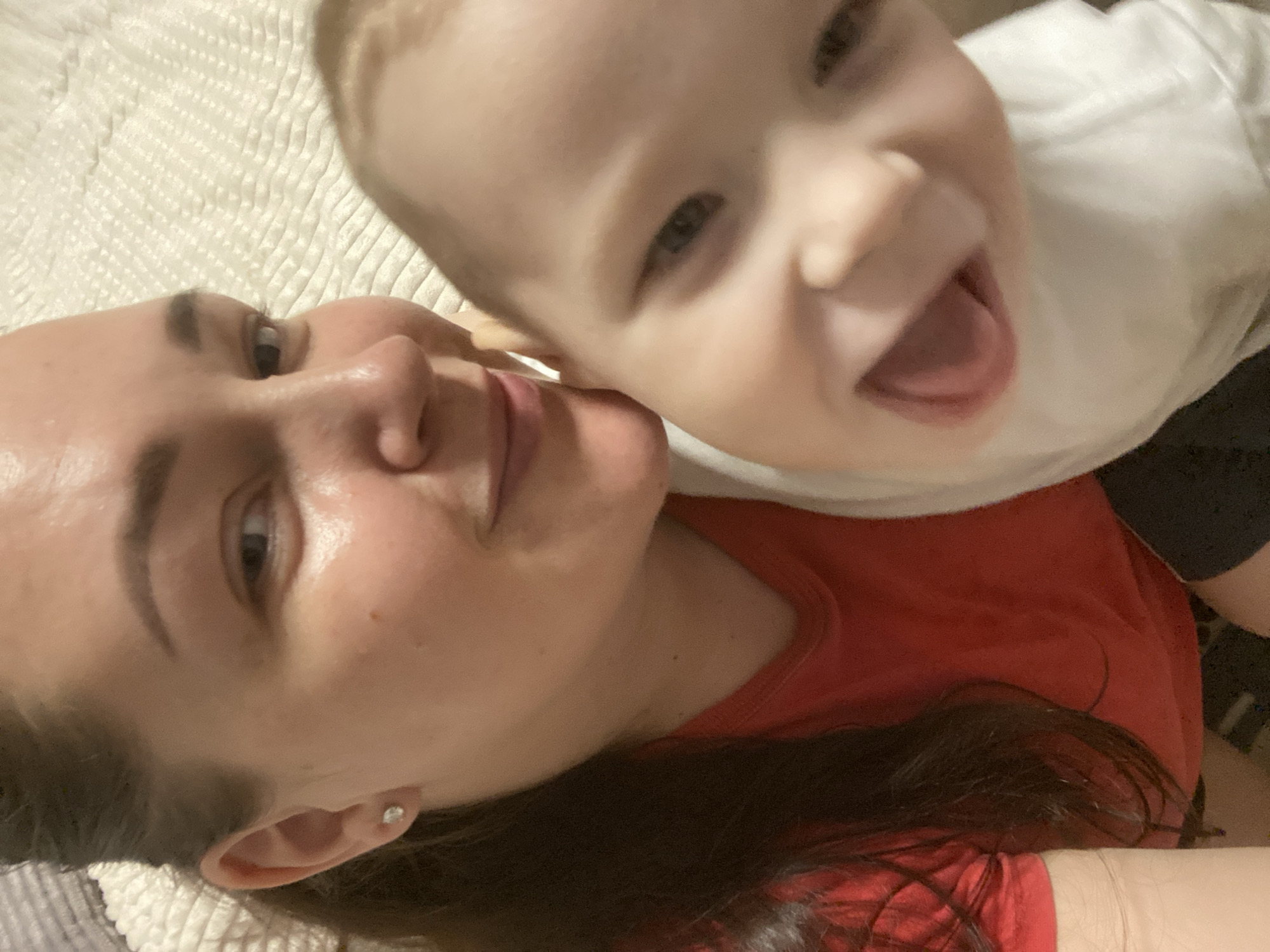 A close-up selfie shows a woman and a baby lying on a textured, possibly quilted surface. The woman has dark hair and is wearing a red shirt. She is looking at the camera with a slight smile. The baby, wearing a white shirt and dark bottoms, is positioned above her, also looking at the camera with an open mouth and a broad smile, sticking out their tongue.
There is no evidence from the image to infer the location of Sochi, Russia.