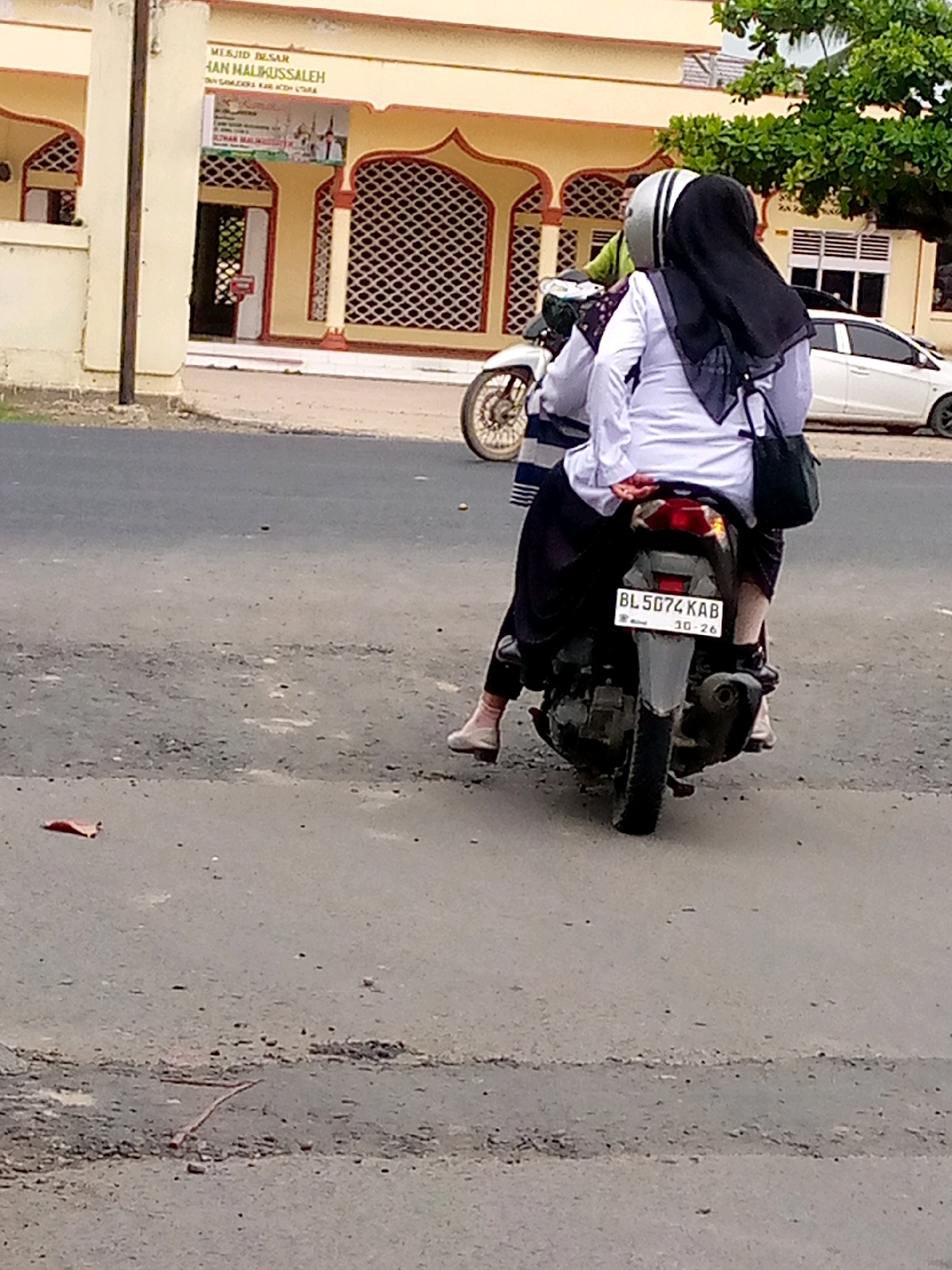 In Keudegeudong, Indonesia, two women are riding on a motorcycle in front of a mosque. The woman in the front is wearing a white shirt, a black hijab, and a black bag slung over her shoulder. The woman in the back is wearing a white shirt, a black hijab, and has her arm around the front rider. Both women are wearing light-colored shoes and dark-colored pants. The motorcycle has a license plate that reads "BL 5074 KAB 10-26". The mosque in the background is yellow with red trim and has a large arched entrance. A white car is parked to the right of the mosque. The ground is paved with asphalt and has some debris scattered on it.