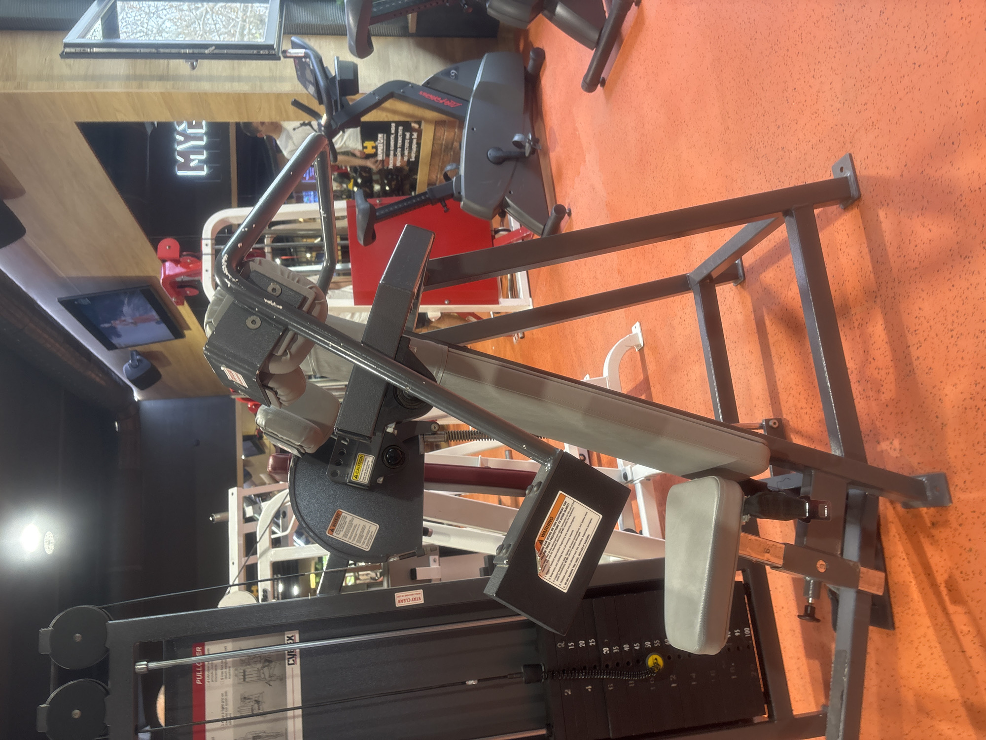 This media file captures an indoor gym setting in Plovdiv, Bulgaria, focusing on various exercise equipment. The scene features a prominent pulldown machine in the foreground, characterized by its black frame, grey padded seat and chest rest, and a weight stack with visible numbers. Warning labels are affixed to parts of this machine, and a manual or instruction sticker reads "PULLOVER" and "CVE EX," likely indicating the brand Cybex. The weight stack shows increments like "20, 25, 30, 35, 40, 45, 50, 55" and pin selections "2, 4, 6, 8, 10, 12." The text "STAY CLEAR" is also visible.
In the background, other gym equipment is present, including a stationary bike identifiable by the "Life Fitness" logo. A red machine and various metal structures of other fitness apparatus are also seen. The gym has distinct orange, textured walls, contrasted by wooden paneling and dark, reflective surfaces, possibly mirrors. A large window offers a glimpse of tree branches outside, suggesting it is daytime. A light fixture or screen displays "MYE," and another screen is visible lower down. A reflection of a person in a white shirt can be seen in one of the reflective surfaces. Other visible text includes "HAMMER GYM" and "тежестите" (Bulgarian for "the weights"). No specific activity is depicted; the equipment appears ready for use.