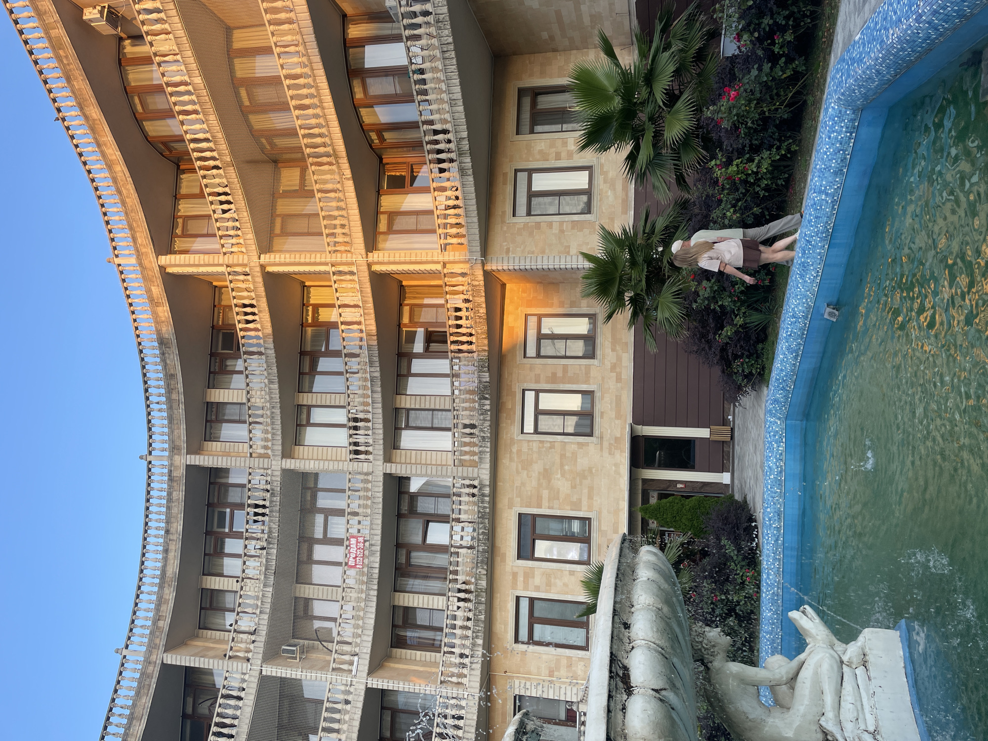 The image depicts a curved building with multiple balconies, likely an apartment complex or hotel, in Sochi, Russia. In the foreground, a fountain with a decorative sculpture is partially visible, with its blue tiled basin filled with water. Two individuals, a woman and a man, are walking away from the viewer alongside the fountain. The building's facade is made of light-colored brick, and it features numerous windows and balconies with ornate railings. The scene is bathed in warm, golden light, suggesting it was taken during either sunrise or sunset. Lush green vegetation, including palm trees and flowering bushes, surrounds the fountain and the building. A red and white "FOR SALE" sign is visible on one of the balconies.
