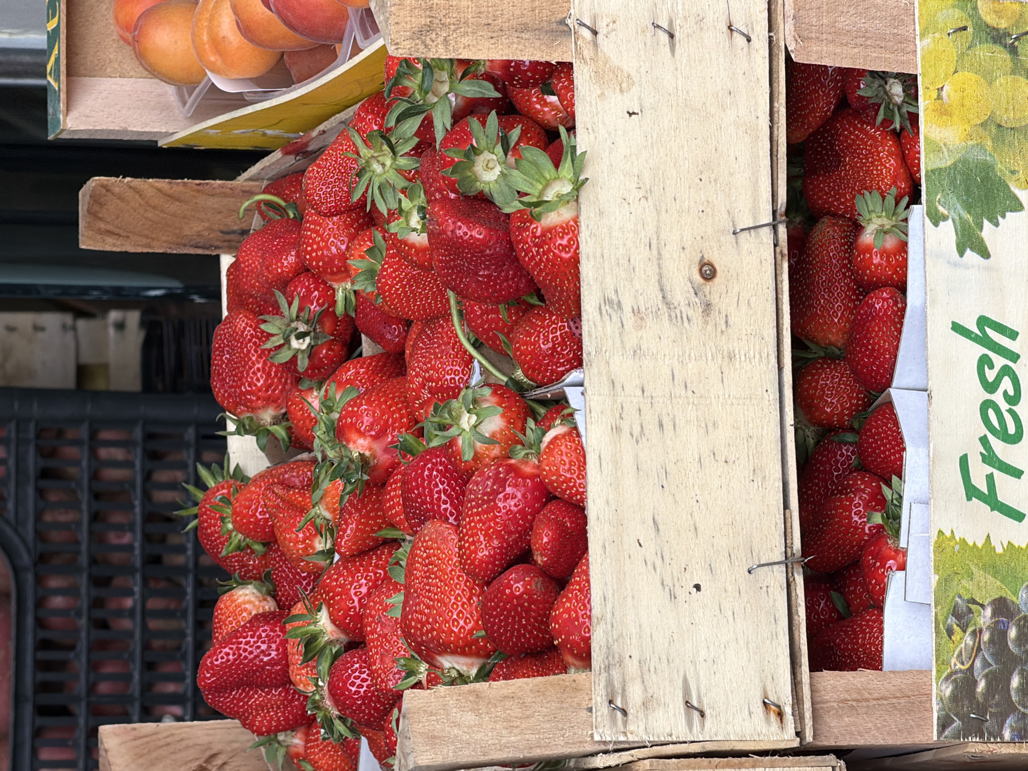 A close-up captures a box of fresh strawberries on display, likely for sale, in Donji Pobori, Montenegro. The strawberries are bright red and piled high within a wooden crate, their green stems visible. Adjacent to the strawberry box is a crate of apricots. A "Fresh" label with an image of grapes is partially visible on the side of one of the boxes. A black, slatted crate is visible in the background. The image is shot in daylight.