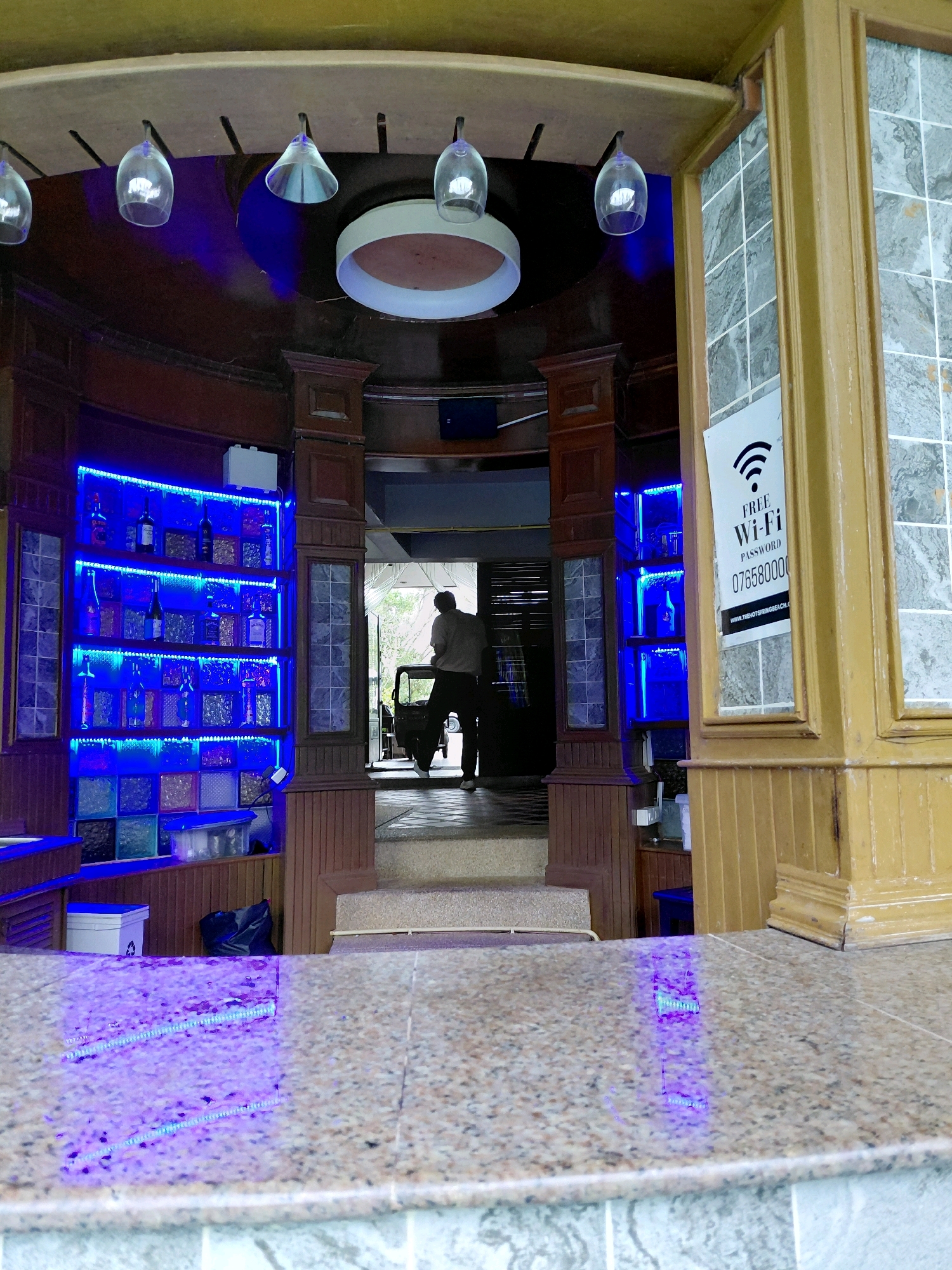 The image captures an interior view of what appears to be a bar or cafe in Ban Khuan, Thailand. The foreground features a polished granite countertop, reflecting purple and blue lights. Behind the counter, shelves are illuminated with vibrant blue LED lighting, displaying various bottles and glass blocks.
To the right of the shelves, a wooden pillar supports a wall tiled with grey and white squares. A white sign with a Wi-Fi symbol, the text "FREE Wi-Fi," "PASSWORD 0765800000," and a website address is attached to this pillar.
In the background, a doorway leads to an outdoor area. A person is silhouetted, standing near a three-wheeled vehicle, possibly a tuk-tuk, which is positioned in the doorway. The lighting suggests it is either late afternoon or early evening. Several wine glasses are hanging upside down from a fixture above the shelves. The overall ambiance is dimly lit with a prominent use of blue and purple lighting.