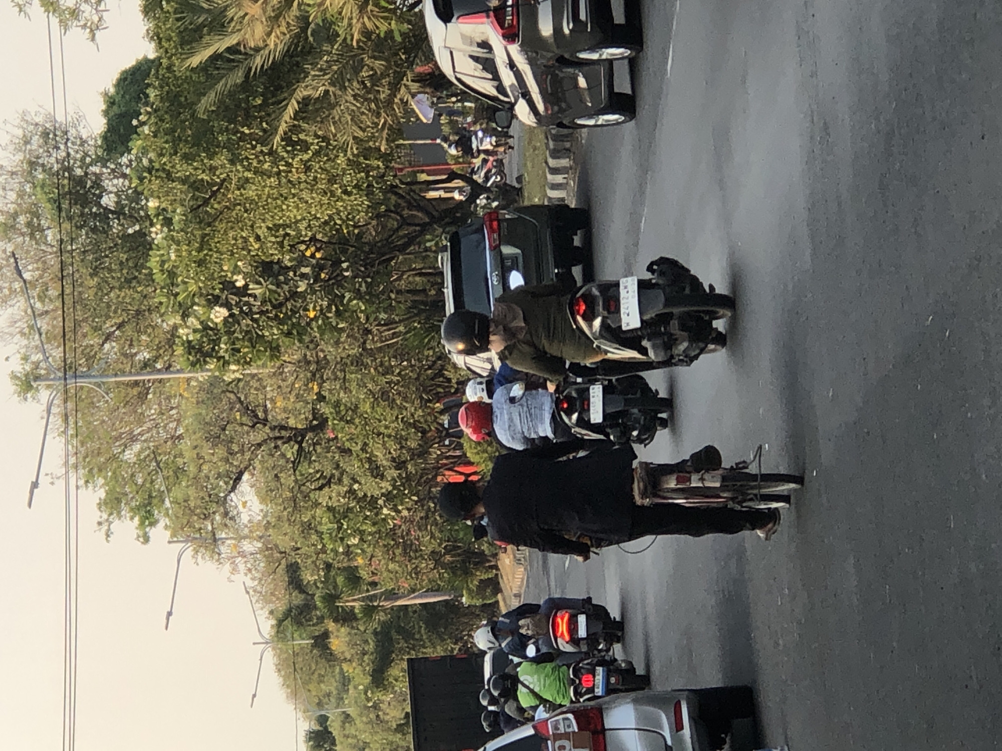 The image depicts a street scene in Ngaban, Indonesia, with several vehicles and pedestrians. In the foreground, a person wearing a black suit is riding a bicycle. Behind this cyclist, a person in a green shirt is riding a scooter, followed by another scooter with a red helmet on the rider. Further back, a dark-colored SUV is visible, and to the right, a dark-colored car is on the road. The environment includes trees with green foliage and some flowering trees. The sky is overcast. The road appears to be asphalt. Streetlights are visible in the background.