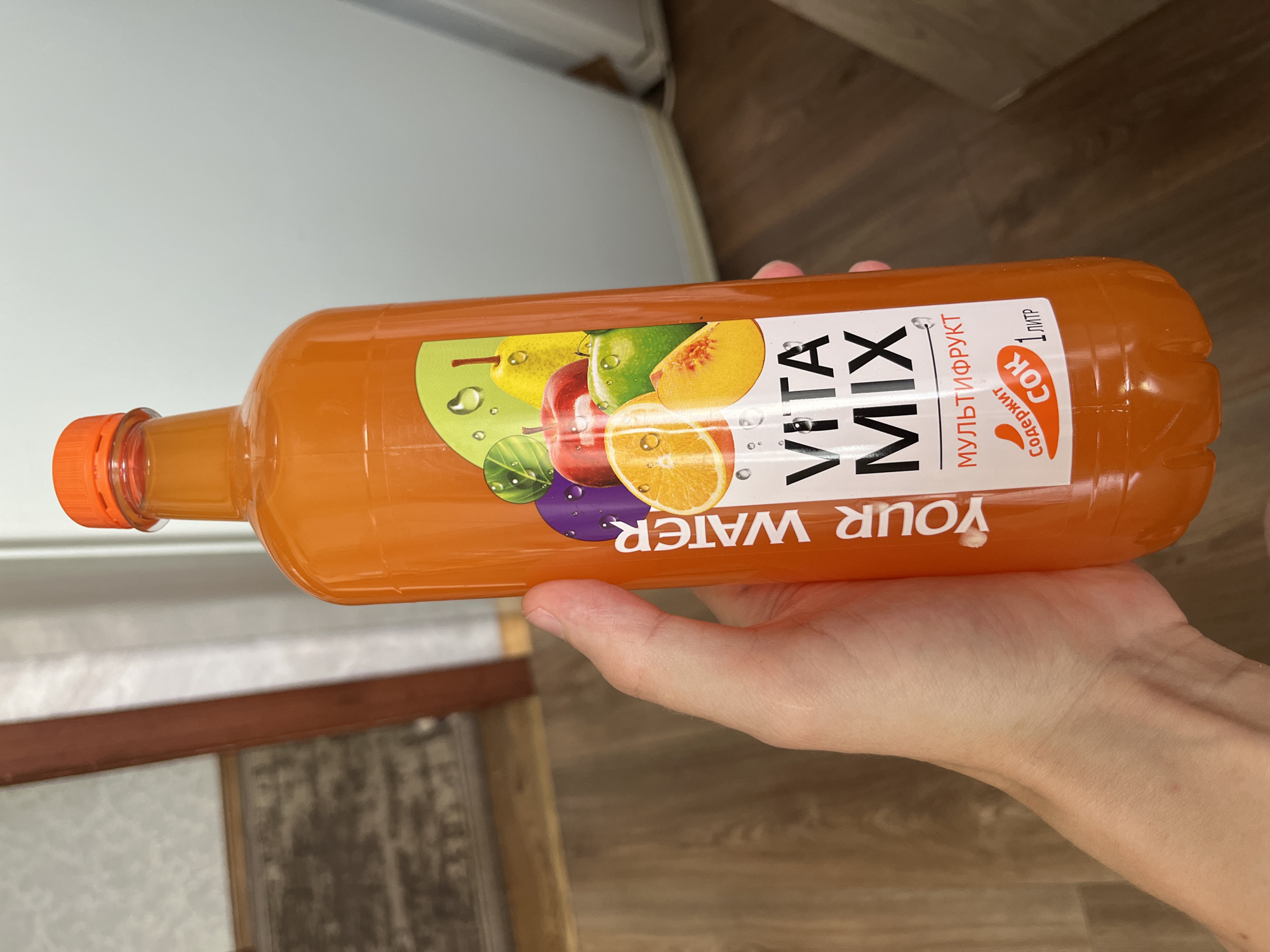 The media file is a photograph of a hand holding an orange plastic bottle of juice. The bottle has a white label with colorful illustrations of fruits such as an apple, pear, peach, orange, and plum. Text on the label reads "YOUR WATER VITA MIX" in large black letters. There is also text in Russian which translates to "MULTIFRUIT" and "JUICE 1 LITER". The bottle is filled with an orange-colored liquid. The background shows a wooden floor and a white surface, possibly a countertop or wall. The lighting suggests it is daytime. The scene appears to be indoors.