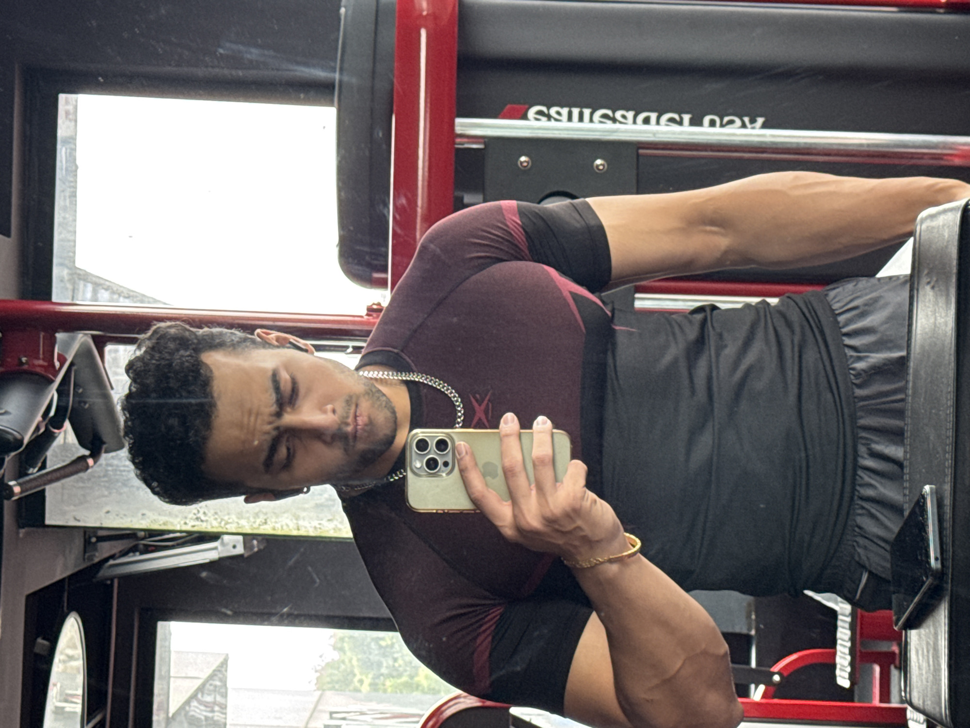 A young Indian man, with dark curly hair and a light beard, stands in a gym in Kolkata, India. He is wearing a maroon and black t-shirt, grey shorts, and a silver chain. The man is holding a smartphone in his right hand and appears to be taking a selfie. His left arm is bent at the elbow, showcasing his bicep. The gym equipment is visible in the background, including a red and black weight machine. A mobile phone is lying on a black surface in the foreground.