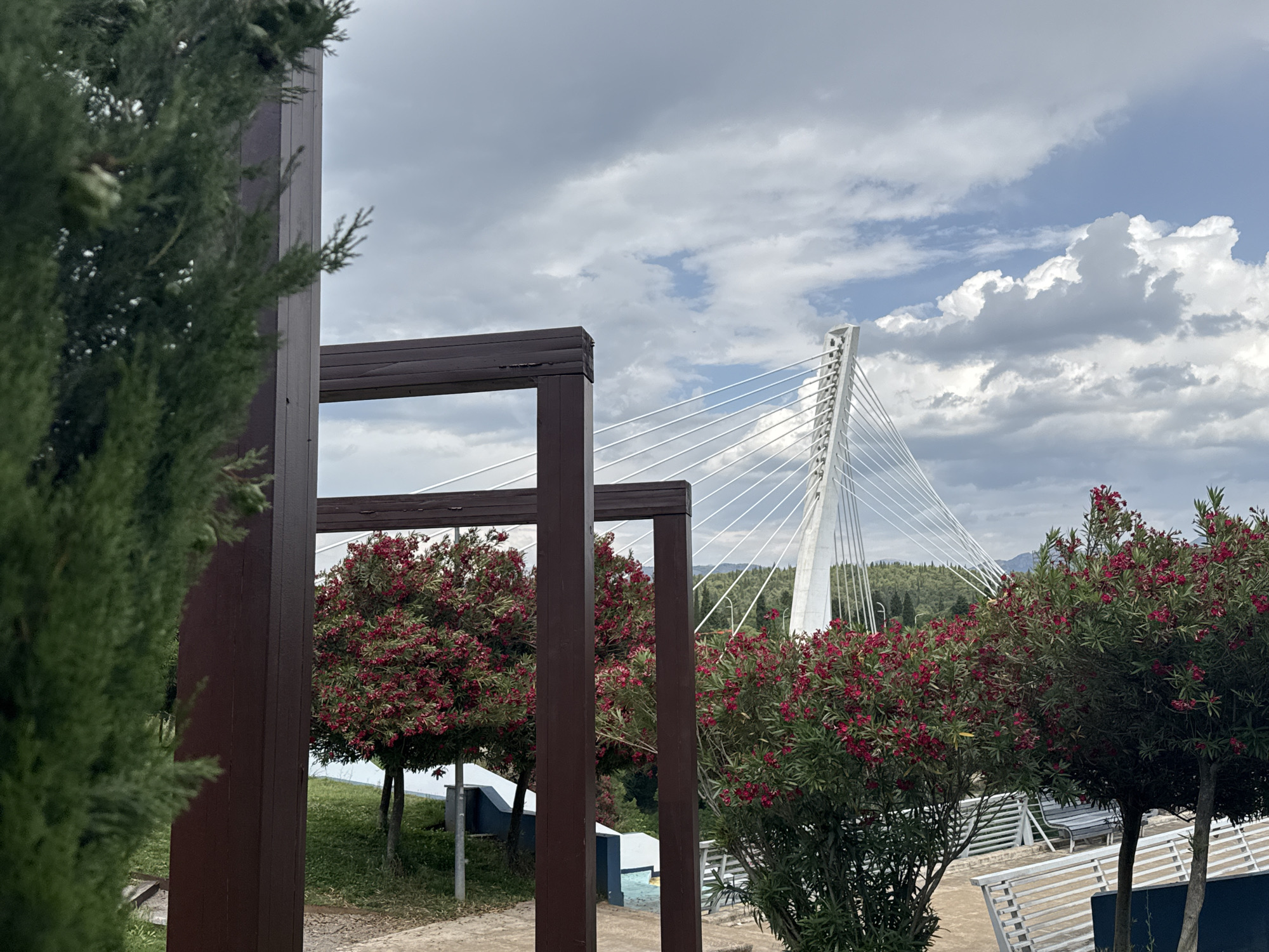 The image depicts a view of the Millennium Bridge in Podgorica, Montenegro, framed by wooden structures and greenery. In the foreground, there are dark brown wooden frames, possibly part of a pergola or similar structure. A tall, slender evergreen tree stands to the left, partially obscuring the view. Between the frames, there are round trees with red flowers and green leaves. To the right of the frames, more of these flowering trees are visible, along with a partially visible white bench. The Millennium Bridge, a modern cable-stayed bridge, dominates the background. Its white, angled pylon and the numerous cables extending from it are prominent. The sky is partly cloudy, with patches of blue visible between the clouds. Distant hills and trees are faintly visible behind the bridge. The scene appears to be taken from a slightly elevated position, with a path or walkway visible in the lower-left area.