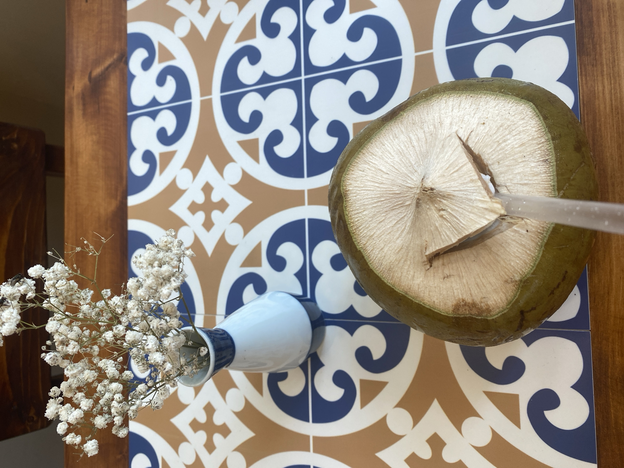 An overhead shot captures a halved young green coconut with a straw inserted, positioned on a table with a patterned tile surface. The coconut's fibrous interior is visible, with a distinct triangular cut at the top where the straw enters. To the left of the coconut, a small, white ceramic vase holds dried baby's breath flowers. The table itself has a warm wooden edge framing the tiled surface. The tile pattern consists of brown, white, and blue geometric shapes. The scene suggests a casual setting, likely a cafe or restaurant in Đà Nẵng, Vietnam, given the vibrant tile design often seen in the region. There are no people or animals visible in the image.
