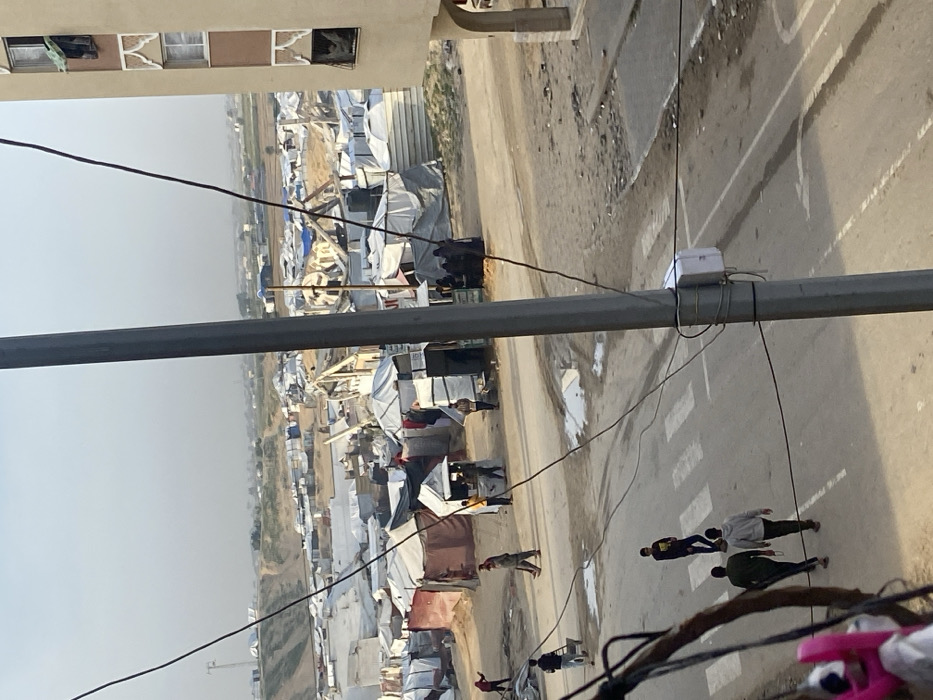 The image depicts an elevated view of a densely populated, informal settlement or camp area in Khan Yunis, Palestinian Territory, adjacent to a multi-story building and a street.
In the foreground, a grey utility pole, adorned with numerous black electrical wires and a white junction box, occupies the central-left. A multi-story beige building with rectangular windows, two featuring decorative arched top frames, stands on the right edge. Laundry hangs from one window.
Below, a paved road with white zebra crossing stripes is visible, showing several wet patches or puddles. On this road, five individuals are present: two men walk away from the camera, one man walks towards the camera on the zebra crossing, another pushes a wheelbarrow on the far left, and one walks horizontally across the street further back.
The mid-ground is dominated by a sprawling, informal settlement comprising numerous makeshift shelters and tents, predominantly constructed from white/light grey tarpaulins and darker fabric materials. Several black cylindrical water tanks are visible on or beside these structures. Small, open-front market-like stalls are interspersed among the shelters, with groups of people, including women in dark attire, gathered near them. In the central-right mid-ground, extensive debris and partially collapsed structures, characterized by exposed wooden beams and twisted materials, are evident, indicating significant damage.
The background reveals a barren, undulating landscape with prominent earthen mounds or berms, extending under a hazy, light blue sky. Further scattered structures are visible in the distance.