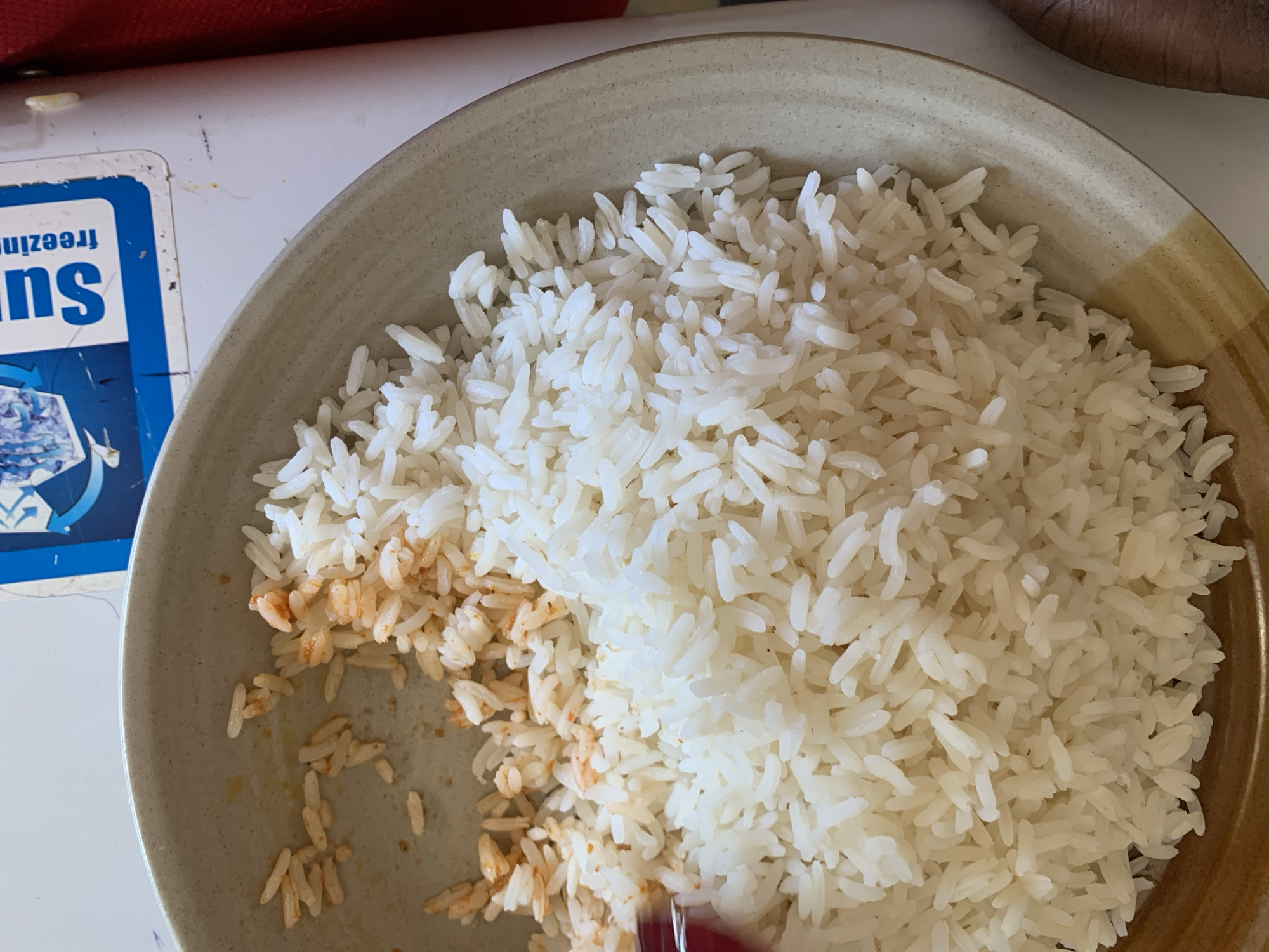 The image provides an overhead, close-up view of a light beige ceramic bowl containing cooked white rice. The majority of the rice appears fluffy and plain white. In the lower-left section of the bowl, some of the rice is mixed with a reddish-orange sauce or seasoning, suggesting it has either been stirred or previously held another food item. The bowl has a subtly speckled interior and a two-toned rim, featuring a lighter beige band adjacent to a darker brown band.
The bowl is placed on a light-colored surface, likely a table or countertop. To the left of the bowl, a blue and white object, possibly a label or package, is partially visible and appears inverted. On this object, the word "Suns" is clearly legible in white on a blue background, and the word "freezing" is partially visible upside down below it. A small portion of a red object, possibly a utensil handle, is visible at the very bottom right edge of the frame.
The scene captures a simple meal of rice, with an apparent eating activity suggested by the mixed portion. No people are visible. The provided location context is New Stadium, Nigeria, though no specific visual cues within the image confirm this geographical setting.