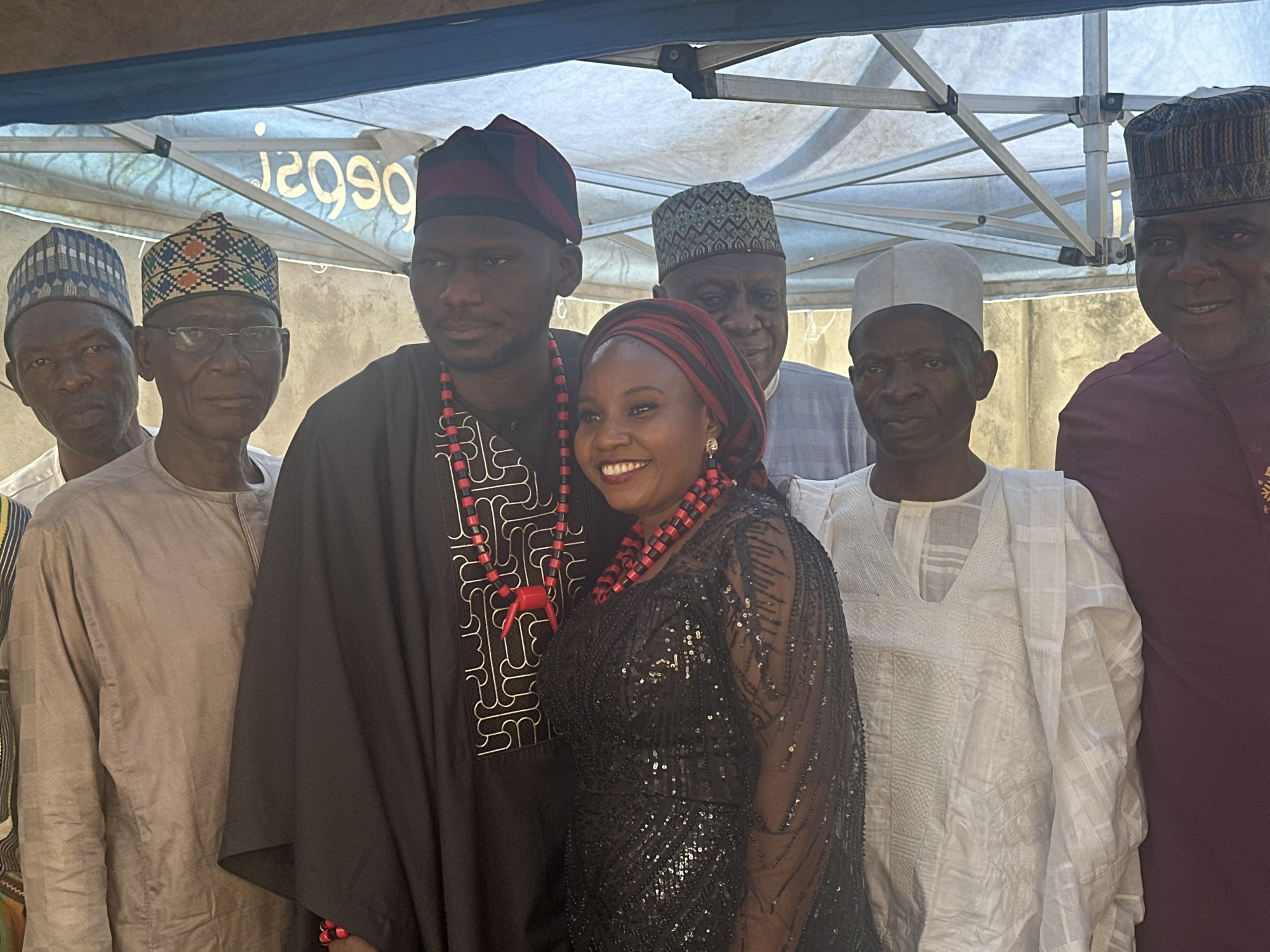 A group of seven adults, predominantly men, is captured in a medium shot, posing closely for a photograph outdoors under a light-colored canopy or tent. The individuals appear to be of African descent and are dressed in traditional Nigerian attire, suggesting a cultural or celebratory event taking place during the daytime.
At the center-right of the frame, a woman stands out with a bright, joyful smile directed at the camera. She is adorned in a black, sequined dress, a red and black striped headscarf, and prominent matching red and black beaded necklaces. Beside her, slightly to the left, is a man wearing a dark, intricately embroidered robe, a dark cap with red stripes, and similar red and black beaded necklaces. He also has a gentle smile.
To the left of the central man, two older men are visible. One wears a multi-colored geometric patterned cap and glasses, while the other, on the far left, wears a blue-patterned cap and a light-colored tunic. On the right side of the woman, an older man in a plain white cap and white tunic is present, and further right, another man in a maroon cap and tunic smiles subtly. A seventh man, wearing a green-patterned cap and a light blue tunic, is partially visible behind the central figures.
The setting includes a light-colored wall or building in the background. The metal frame structure of the canopy is evident above the group. Bright, even lighting indicates it is daytime. On the canopy fabric behind the group, reversed text, likely "PEPSI," is visible. The collective mood appears to be festive and social.