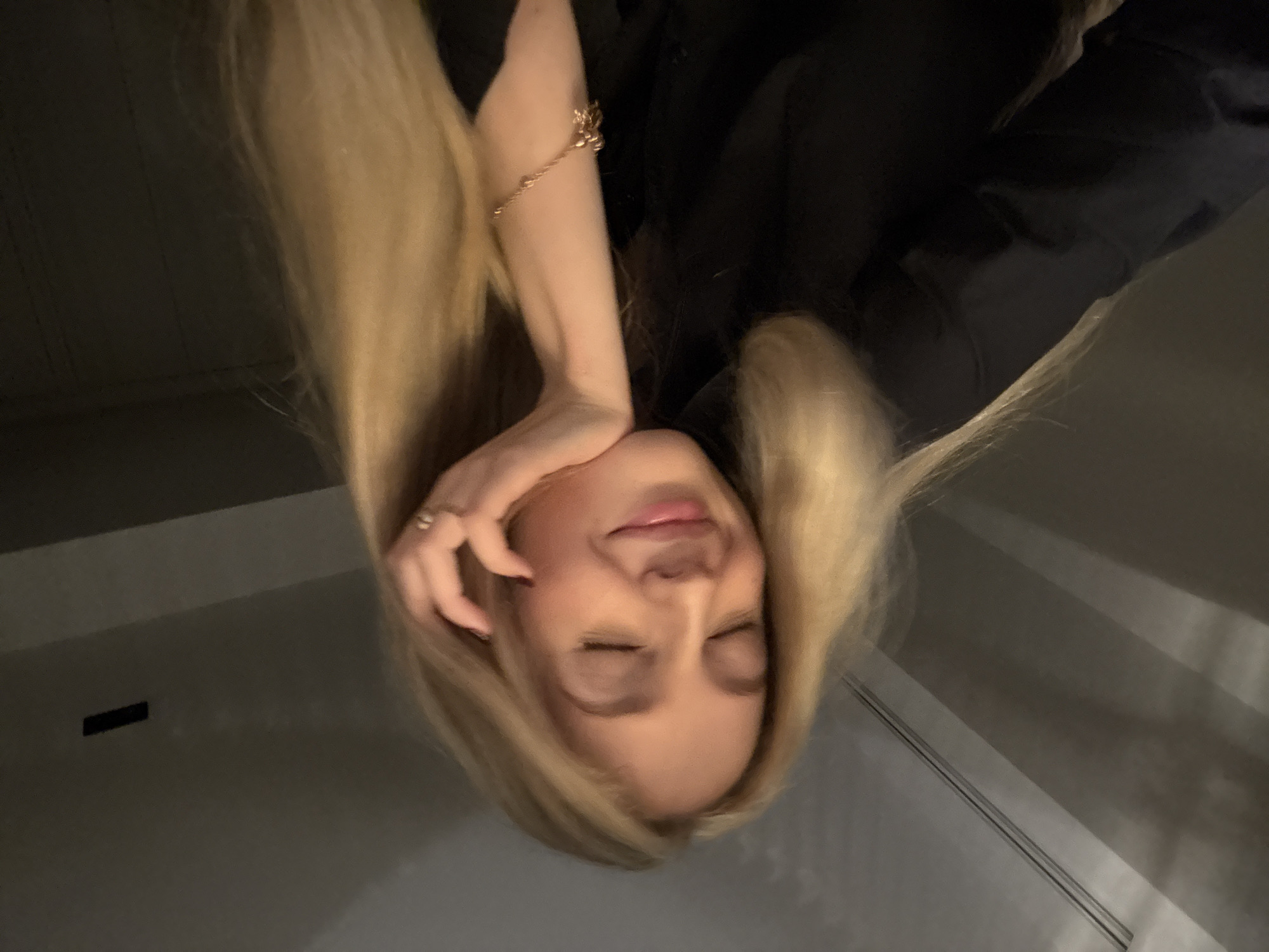 A woman with long blonde hair and closed eyes is photographed at an upside-down angle. She is wearing a black shirt. Her right hand is touching her face, and a gold bracelet is visible on her wrist. A ring is visible on her finger. The background is a neutral gray, with what appears to be a wall and faint shadows. This photograph could have been taken anywhere; there's no indication it was taken in Mexico City.
