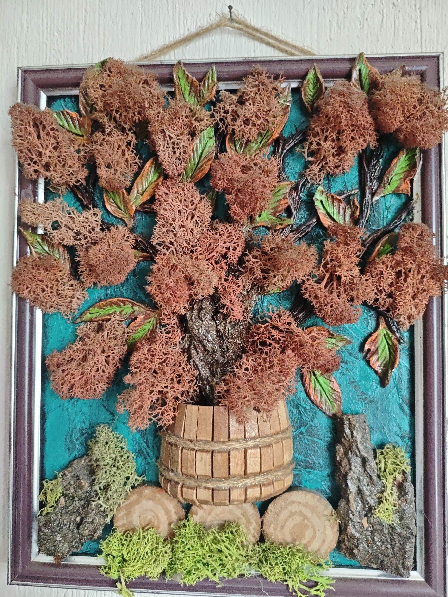 The image shows a framed artwork. The frame is made of brown-silver plastic. Inside the frame, a miniature scene is depicted. The background is a textured turquoise surface. A miniature wooden barrel sits in the center, overflowing with brown, coral-like material that resembles flowers or moss. Stylized leaves, painted in green and brown, are scattered among the "flowers." Dark brown, artificial-looking branches can be seen behind the "flowers." At the bottom of the artwork, there are pieces of tree bark and cross-sections of small branches, surrounded by bright green moss. The artwork is hung by a brown twine. The image does not show any location in the city of Kulata, Bulgaria, nor does it contain information about people, subjects, objects, actions, interactions, and the environment related to that location.