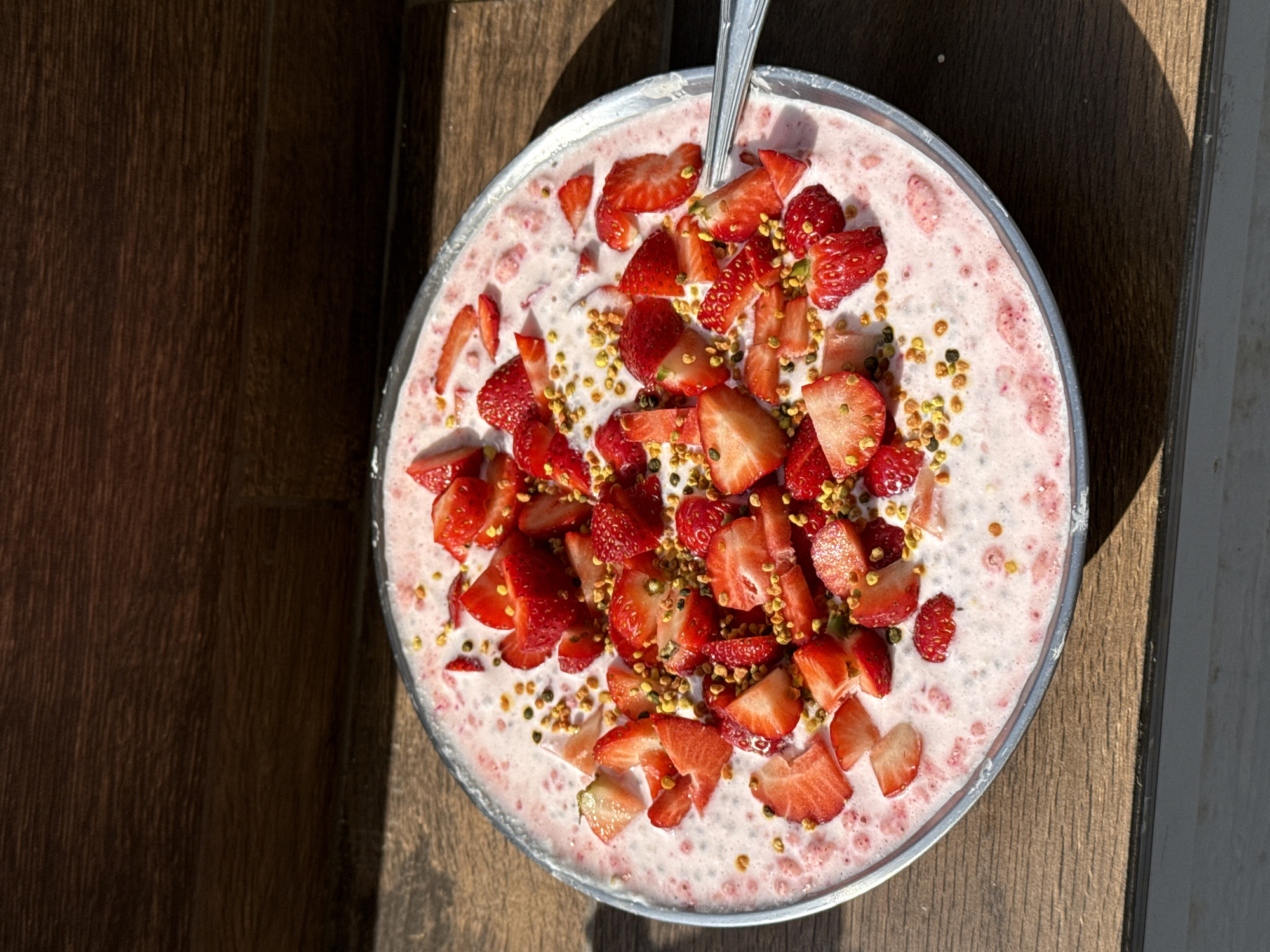A top-down shot shows a metal bowl on a wooden surface. The bowl contains a pink smoothie or yogurt mixture topped with chopped strawberries and small yellow pellets (possibly bee pollen). A spoon is partially visible in the bowl. The wooden surface appears to be part of a table or counter. A bright light is shining from the upper right, casting shadows. No people are visible. There is no indication of the location being Sofia, Bulgaria.