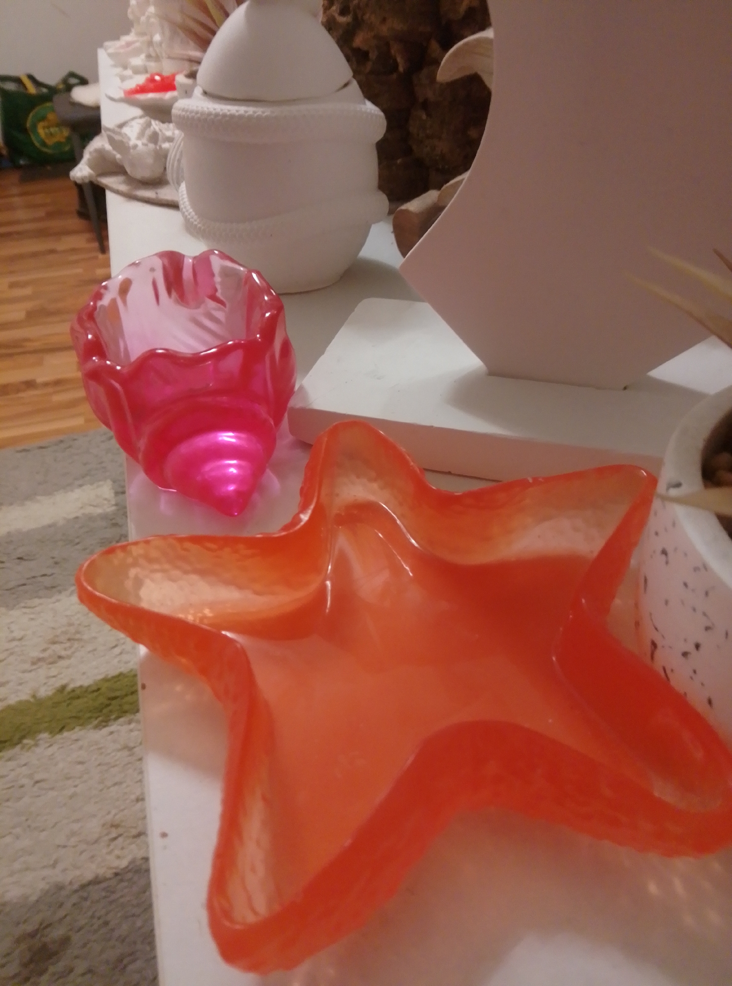 The image displays a collection of decorative objects on a white surface, possibly a table, with a carpeted floor visible in the lower left corner. In the foreground, a bright orange, star-shaped dish is prominent. To its left and slightly behind it is a smaller, pink, shell-shaped dish. In the background, a large white ceramic pot with a textured band around its middle sits on the white surface. Behind the pot, a large, light pink, curved object, possibly a piece of a sculpture or a large decorative item, is visible. Further back, a brown, textured object, possibly dried plant material or bark, is partially obscured. To the far left, a green bag with yellow lettering is leaning against something. The scene appears to be indoors, likely in a residential setting, and does not provide any specific clues to a location in Wedel, Germany.
