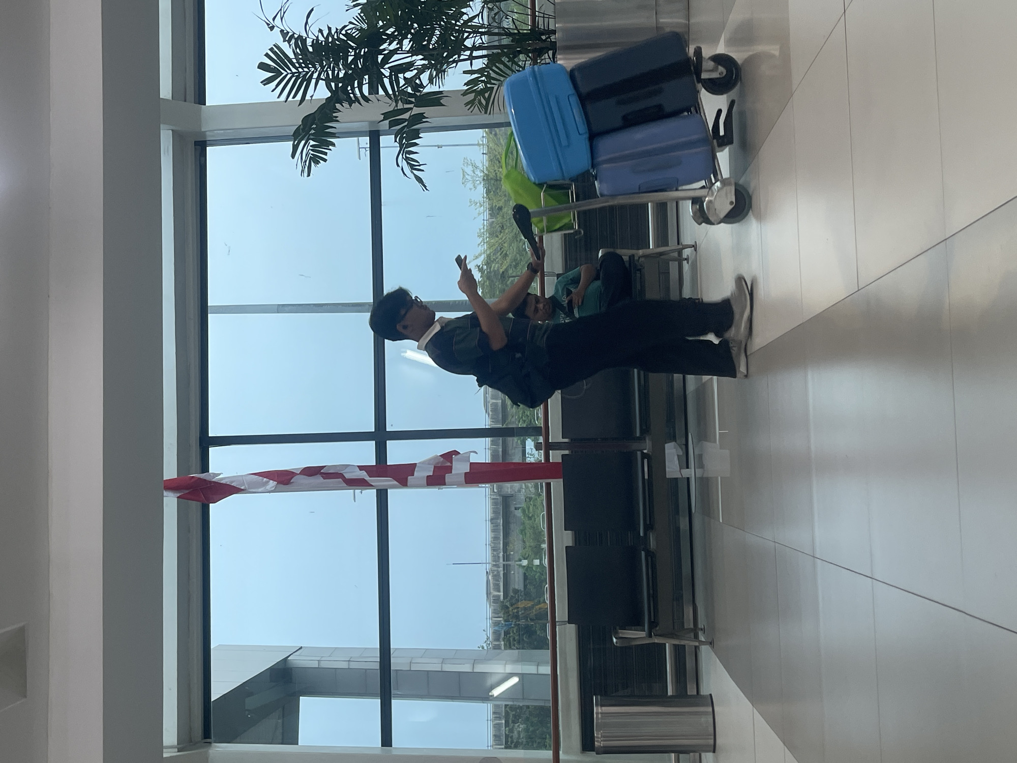 The image depicts a man standing in an airport terminal, holding a smartphone and a luggage cart laden with two suitcases. He is wearing a dark green polo shirt, dark pants, and light gray sneakers. The luggage cart is positioned to his right. In the background, another man is seated, also looking at his phone. Behind them are rows of dark chairs, a large window, and a tall potted plant. A flag with red and white stripes hangs in front of the window, partially obscuring the view of an elevated structure and greenery outside. The floor is tiled with a light gray and white pattern. The overall environment suggests a modern airport waiting area. This scene is observed at Soekarno-Hatta International Airport (CGK) in Tangerang, Indonesia.