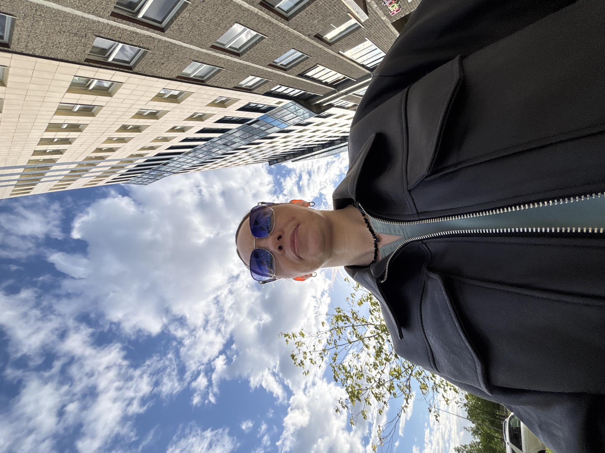 A low-angle shot shows a person wearing sunglasses and a dark jacket standing outside. Above the person is a blue sky with scattered white clouds. In the background, tall buildings with multiple rows of windows can be seen. Green foliage and a white vehicle are visible in the lower right corner of the frame. The location is in the city of Unknown, Unknown.