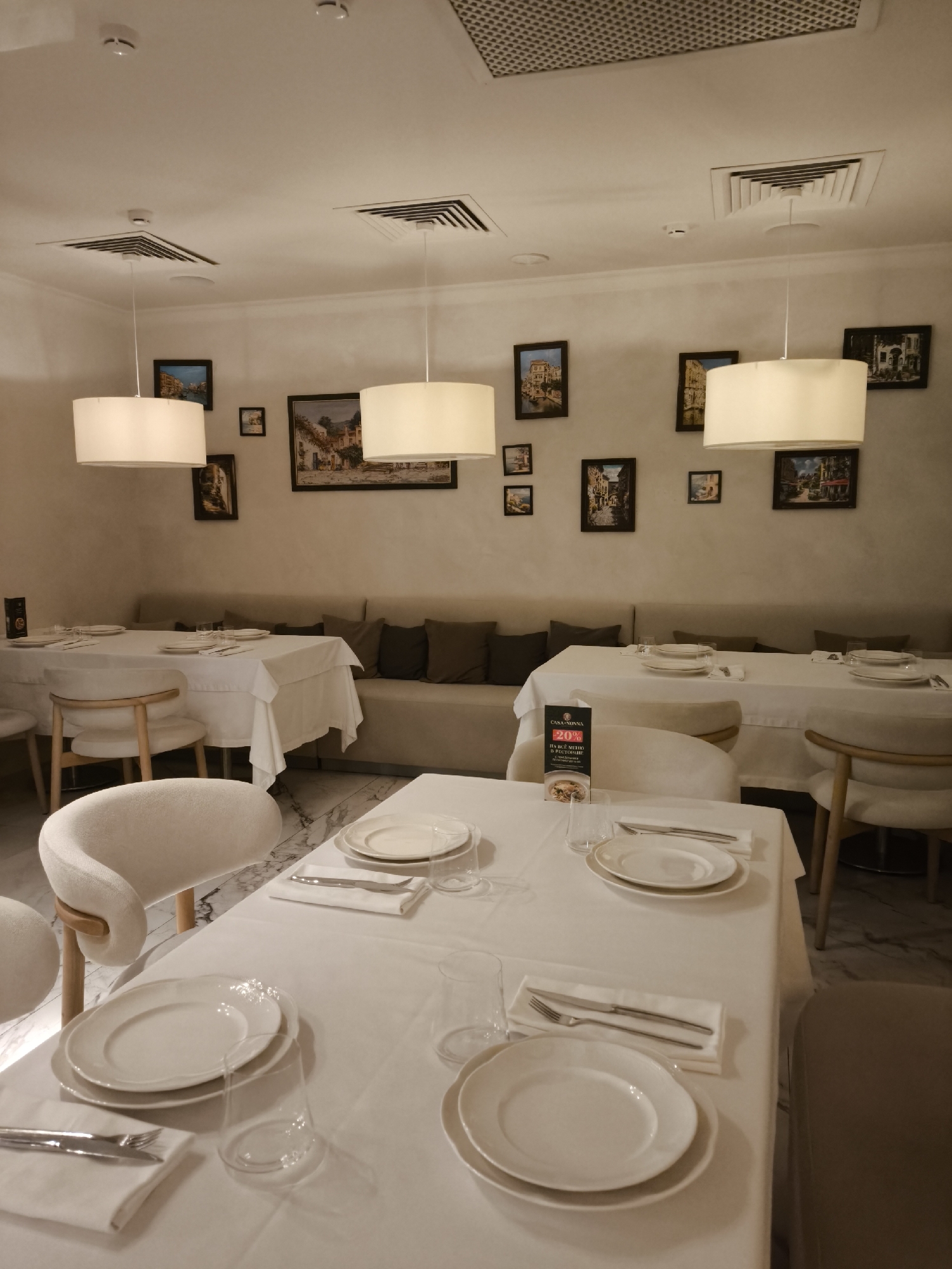 The image depicts the interior of a restaurant in an unknown city. The room is lit with warm, soft light emanating from cylindrical pendant lamps hanging over the tables. The walls are adorned with framed paintings, arranged in a slightly asymmetrical manner. Tables are covered with white tablecloths and set with plates, silverware, napkins, and glassware, indicating a formal dining setting. Several chairs are upholstered in a light-colored fabric and are placed around the tables. In the background, a long, neutral-toned banquette is visible, providing seating along the wall. A promotional sign stands on one of the tables, partially obstructing the view. The floor appears to be marble or a similar polished stone. The scene suggests a sophisticated and intimate dining environment.