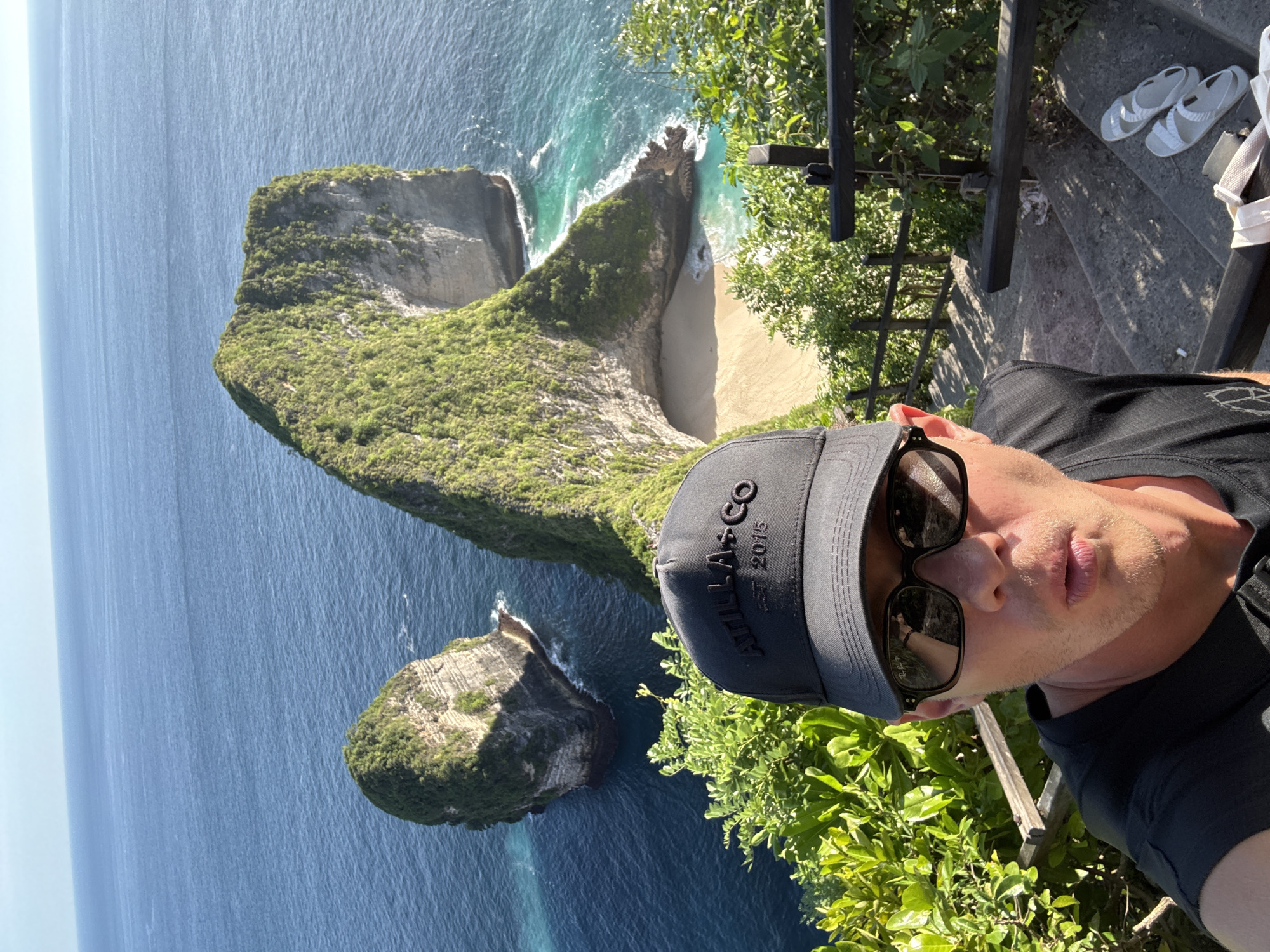 A man is taking a selfie at Kelingking Beach in Nusa Penida, Indonesia. The ocean and the island are in the background. There is a set of stairs to the right of the man with a pair of sandals.