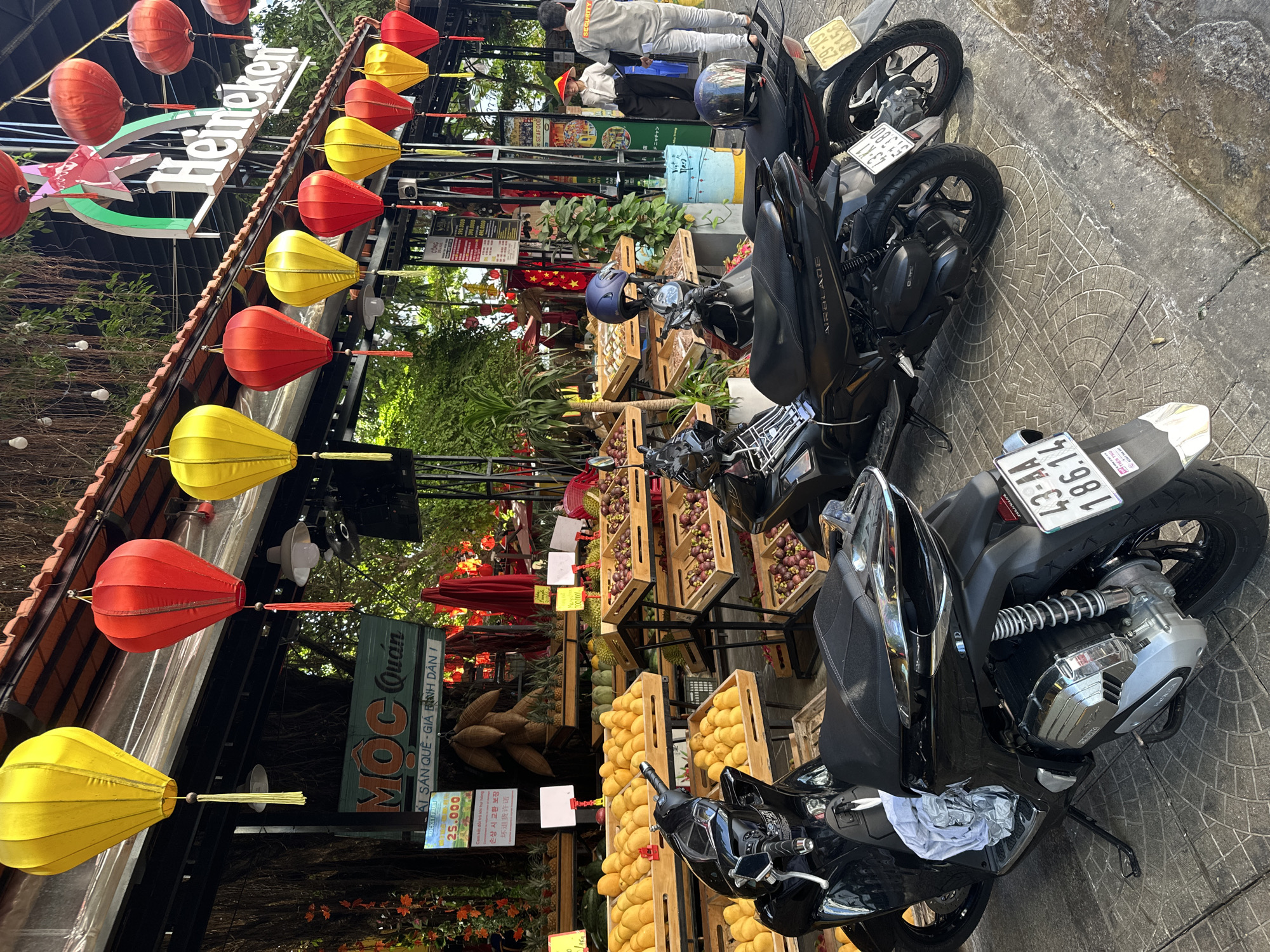 The image depicts a street scene in Đà Nẵng, Vietnam. The scene is oriented at an angle, showcasing a sidewalk and the front of a restaurant or shop. Several black motorbikes are parked on the sidewalk, taking up a significant portion of the space. Alongside the motorbikes, wooden crates filled with fruit (mangoes, mangosteens, durian) are displayed for sale.
The restaurant or shop is adorned with red and yellow lanterns, and a sign that says "MOC Quán" is visible. A "Heileken" (Heineken) sign is visible above. Another smaller sign indicates the price of "ROCKET FRUIT" as 25,000.
In the background, two men, one wearing a grey "SECURITY" shirt, are visible. The overall environment appears to be a bustling commercial area with a mix of street vending and established businesses.