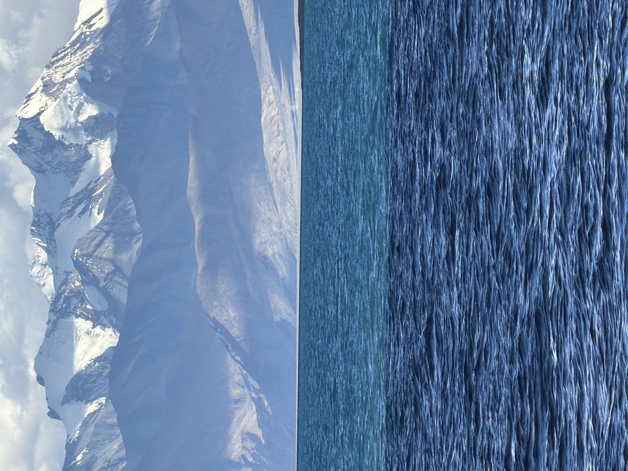 The image depicts a vast expanse of choppy blue water in the foreground, with a distinct line dividing it into two shades of blue. In the middle ground, a low, hazy horizon is visible, and beyond that, a range of majestic, snow-capped mountains dominates the background. The mountains are illuminated by sunlight, highlighting their rugged peaks and slopes. The sky above is partly cloudy, with patches of blue sky interspersed with white clouds. The scene appears to be a natural landscape, likely a lake or a large body of water situated at the foothills of a mountain range. The location in the city of Unknown, India, is not explicitly discernible from the visual information provided.