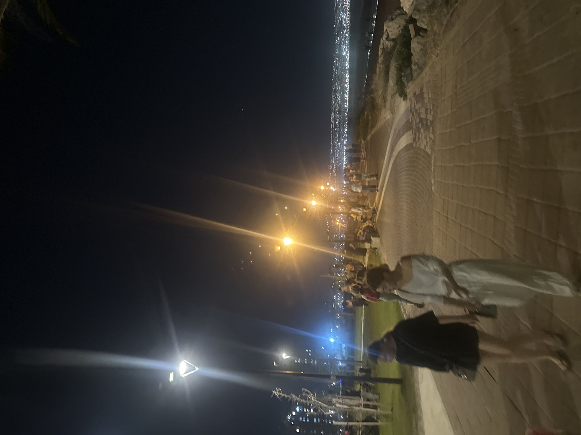 The image depicts a nighttime scene along a waterfront promenade in Kiryat Yam, Israel. A paved walkway, constructed with interlocking bricks in a circular pattern, dominates the foreground. Two women are walking on this path; the woman on the left wears a black top and shorts with a crossbody bag, and the woman on the right wears a white off-the-shoulder top and matching pants, also carrying a bag.
In the background, a park area with green grass and trees is visible, illuminated by streetlights. Further beyond the park, a city skyline with numerous illuminated buildings stretches along the coast. The sea is visible to the right of the promenade, with gentle waves lapping the shore, and rocks are present along the edge of the promenade. The overall atmosphere is that of a public gathering space at night, with people strolling and enjoying the evening. The sky is dark, indicating it is nighttime.
