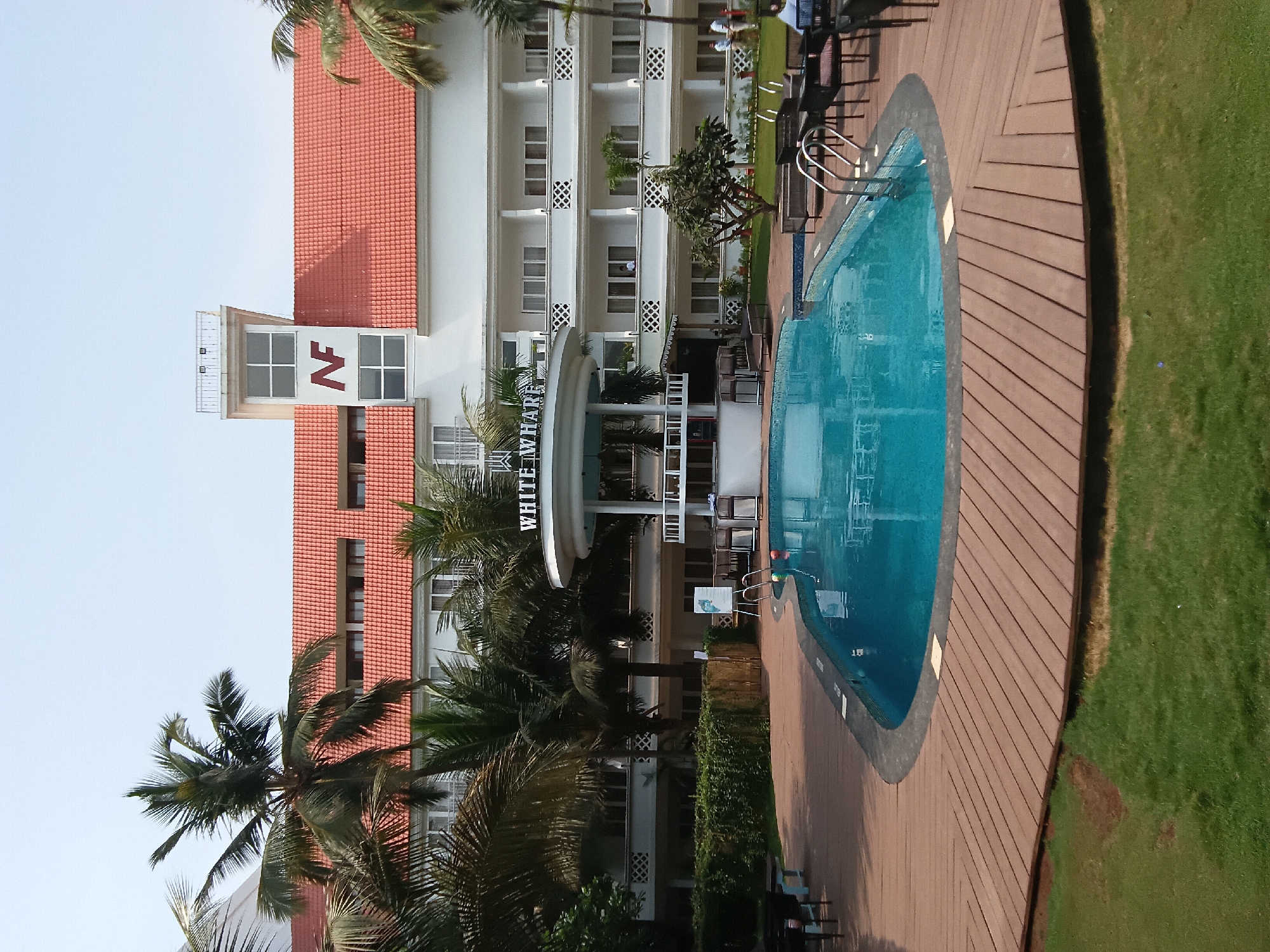 The image shows the White Square Nirvana Hotel in Ashvem, India. The building is white with a red-tiled roof and features balconies. Palm trees are visible in front of the hotel. Next to the hotel is an oval-shaped swimming pool with a wooden deck. A grassy area borders the deck. The sky is clear.