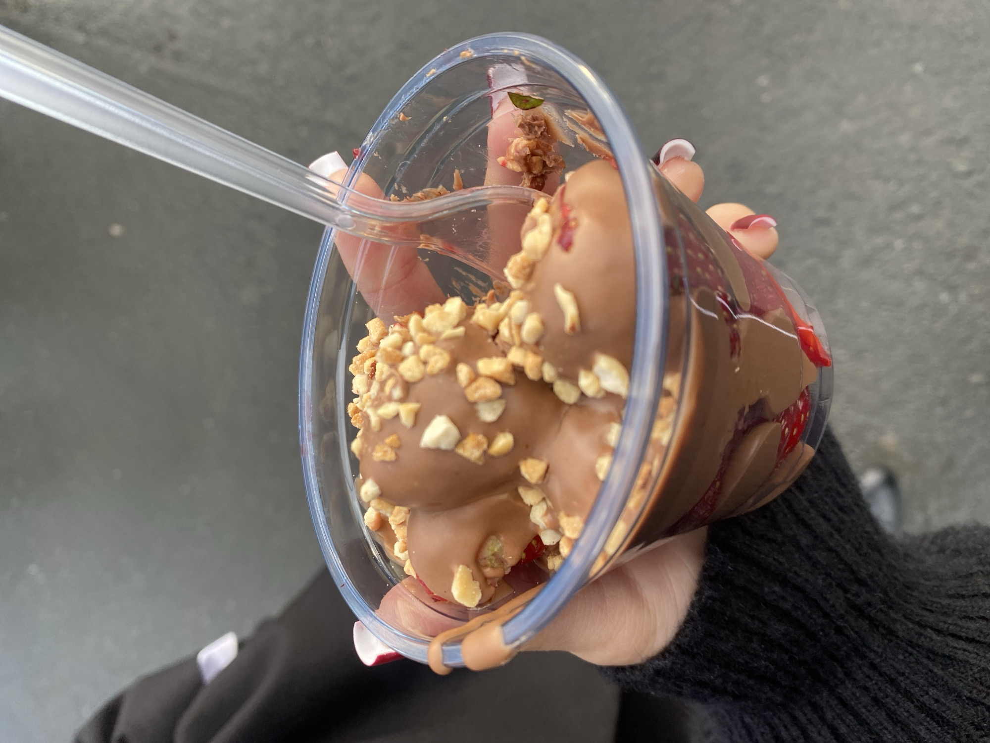 A close-up, eye-level shot shows a person's hands holding a clear plastic cup filled with a dessert. The cup contains chocolate-covered strawberries, some of which are topped with chopped nuts. A clear plastic spoon is partially submerged in the dessert. The background is out of focus and appears to be a gray, textured surface, possibly pavement. The person is wearing a black, knitted sweater. The overall impression is of a casual outdoor setting. The image does not contain any specific landmarks or clues to indicate it was taken in Uchaly, Russia.