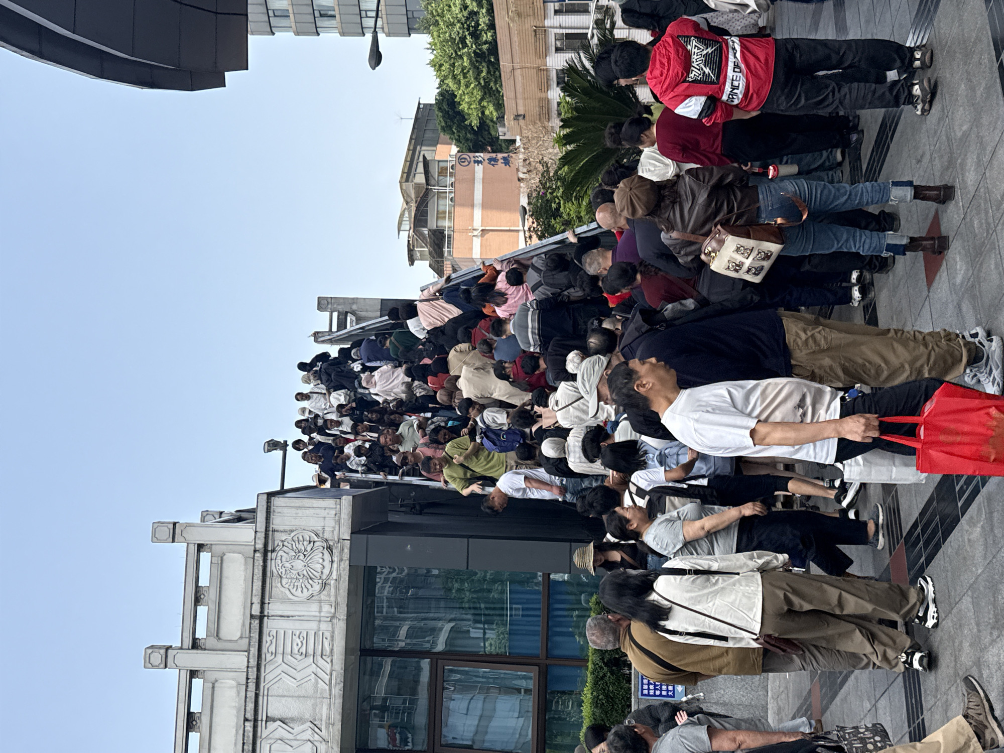The image captures a vast crowd of people, mostly adults, densely packed and queuing outdoors in an urban environment. They are standing on what appears to be a sloped or stepped walkway, extending far into the distance. Most individuals are casually dressed, carrying various bags such as backpacks, handbags, and tote bags, with one person in the foreground holding a bright red shopping bag.
The setting is an urban street or square, characterized by a clear, bright blue sky and ample natural light, suggesting a daytime scene with fair weather. Buildings flank the gathering: to the left, a structure with reflective windows and an ornate, carved facade adorned with traditional Chinese decorative elements is prominent. In the mid-ground, a lighter-colored building displays blue Chinese characters and a circular logo, possibly indicating a financial institution, though specific text is hard to discern. Lush green foliage, including trees and shrubs, is visible behind and between some buildings.
The primary activity is a large-scale gathering or queue, indicative of a significant event, popular attraction, or busy transit point in China, specifically Yuzhong District. The crowd appears orderly, with people patiently waiting. Apart from the potential Chinese characters and logo on the building, some unreadable smaller Chinese characters appear on a blue sign in the lower right.