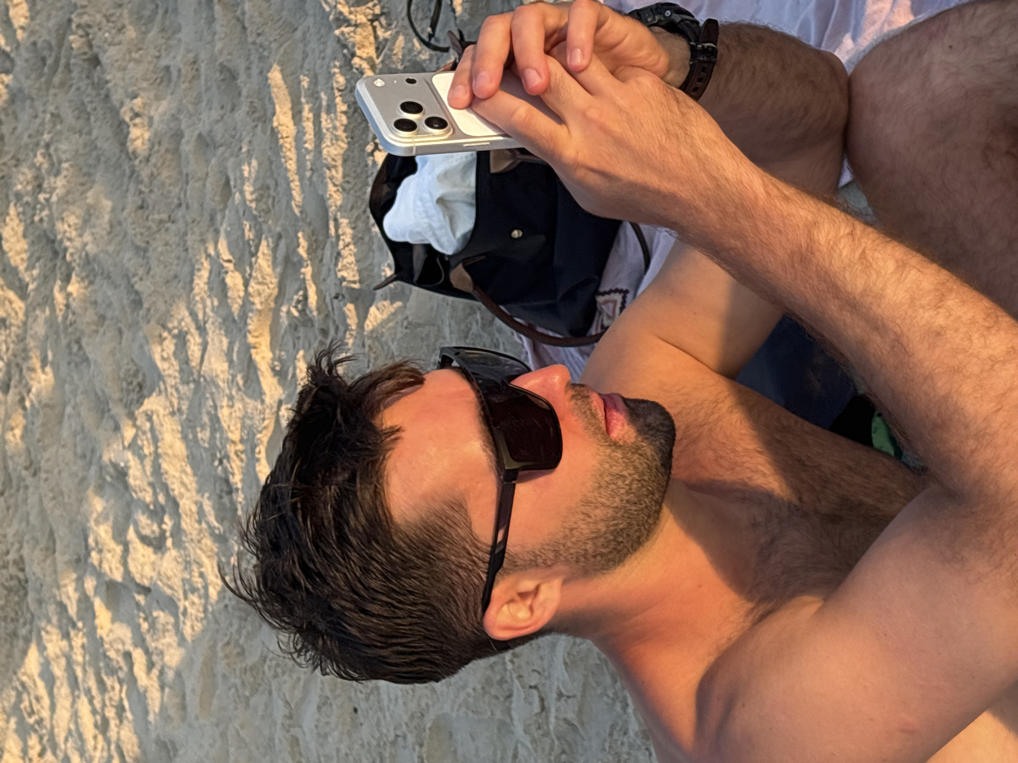 An adult male is depicted seated on a sandy beach in Tel-Aviv, Israel, bathed in the warm, low-angle light of the golden hour. He has short, dark hair and a short beard. He is shirtless, revealing a tanned physique with visible body hair on his chest and arms, which are highlighted by the orange-red hue of the sunlight. He wears dark, rectangular sunglasses. His attention is directed towards a silver/light gray smartphone, identifiable as an iPhone (likely a Pro Max model due to its triple-camera module set on a raised square), which he holds vertically with both hands, his thumbs positioned over the screen. Behind his right arm, resting on a light-colored, patterned towel or blanket, is a dark-colored tote bag with brown leather accents, partially revealing a white garment inside. A dark watch with a textured strap is visible on his left wrist. The immediate foreground and background consist of light brown sand, exhibiting various ripples and indentations.