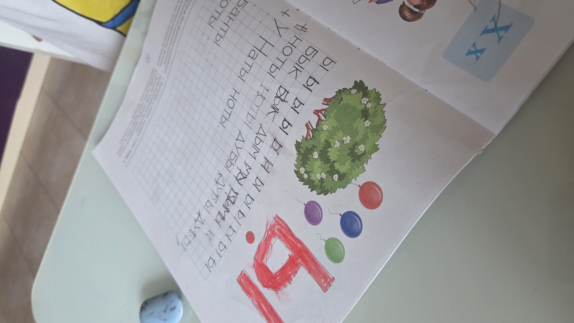A children's workbook is spread open on a light-colored table in what appears to be an indoor setting in Chaikovsky, Russia. The left page is a grid-lined exercise sheet filled with handwritten Cyrillic letters and words, indicating a writing practice activity.
Visible handwritten Cyrillic text includes repetitions of the letter "Ы", and words such as "БЫК" (Bull), "ДЫМ" (Smoke), "Дом" (Home), "Наты" (Naty's), "НОТЫ" (Notes), "ДУБЫ" (Oaks), and "Банты" (Bows). A large, roughly colored red Cyrillic letter "Р" is prominently drawn at the bottom of this page.
The bottom right portion of the left page and the right page feature colorful illustrations. A green bush with small white flowers is depicted, extending across the page fold. Three balloons in purple, blue, and green colors float near the bottom right. On the top right page, a blue tag with the letters "XX" is visible, along with part of an illustration showing the head and shoulders of a person, possibly a child with brown hair and a purple top. A light blue eraser is resting on the table in the bottom left corner of the frame. A yellow and blue object, possibly fabric or a toy, is partially visible at the top left. The scene depicts a child's educational or homework session. The lighting suggests an indoor setting during daytime.