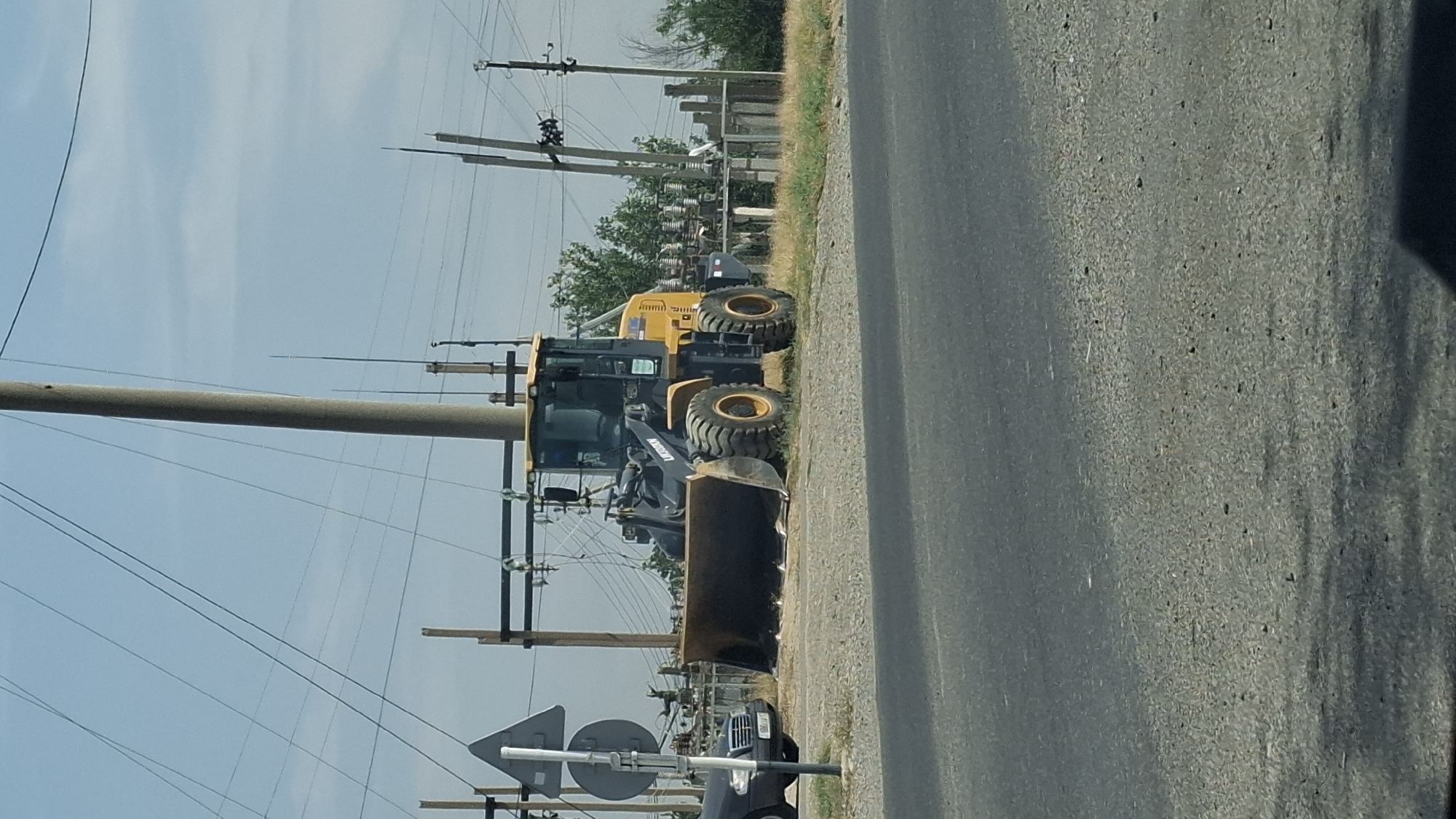 A yellow and black front-end loader is parked on a gravel surface next to a paved road. The loader has a large bucket at the front. In the background, there are utility poles with wires, electrical insulators, and a line of trees. To the left of the loader, a dark-colored car is parked. A traffic sign with a white triangle and a circular sign is visible to the left of the loader. The scene appears to be in an outdoor, possibly rural or semi-urban, area with clear skies. The location is identified as Қасымбек датқа, Kazakhstan.