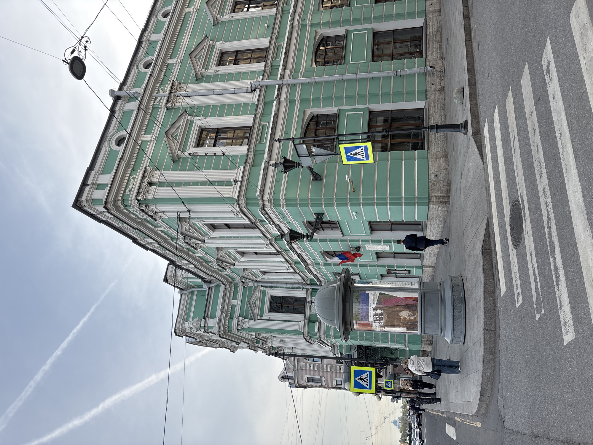 The image captures a street scene in Saint Petersburg, Russia, featuring a prominent green building with white architectural details. The building has multiple windows and ornate trim. A pedestrian crosswalk is visible on the right side of the frame. A cylindrical advertising column stands on the sidewalk. Several people are walking on the sidewalk, and traffic is visible in the distance. A blue and yellow pedestrian crossing sign is attached to a lamppost. A Russian flag appears to be affixed to the building. Contrails are visible in the sky.