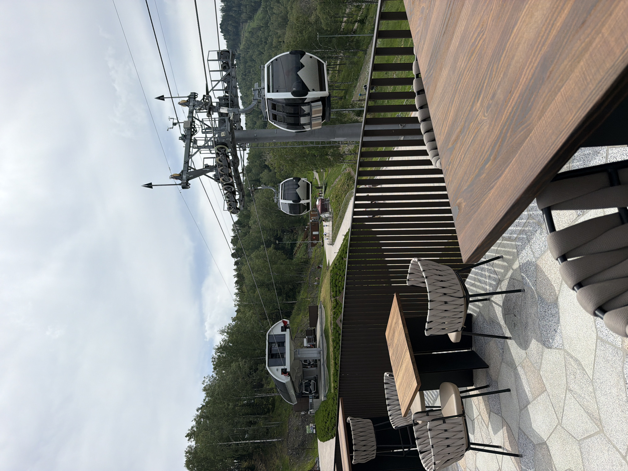 The image shows an outdoor seating area on a paved terrace with a wooden railing. There are several tables and chairs, including bar stools. In the background, a ski lift system with gondolas is visible, with a station and wooded hills beyond. The sky is overcast. The location is identified as Озерное (майминский), Russia.