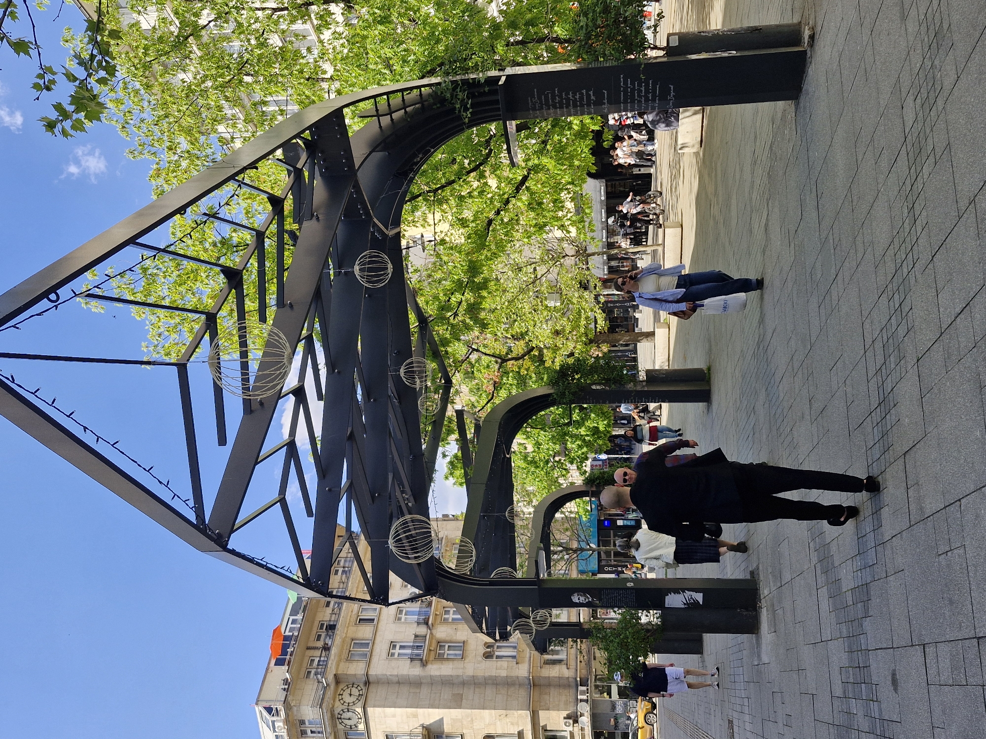 The image shows a pedestrian street in Sofia, Bulgaria, lined with metal archways decorated with globe-shaped ornaments and strings of lights. People are walking along the gray paved walkway. A bald man in a dark suit and shoes, carrying a black bag, is walking towards the camera. Another person is walking in the same direction, slightly behind the man in the suit. Further down the walkway, a man in a blue shirt and jeans walks in the opposite direction, carrying a white bag. Several people are sitting at outdoor tables in the background. Buildings with classical architectural features and visible clocks are on the left side of the image, partly obscured by the archways. Green trees and a partly cloudy blue sky are visible above. Black plaques displaying text are positioned along the walkway.