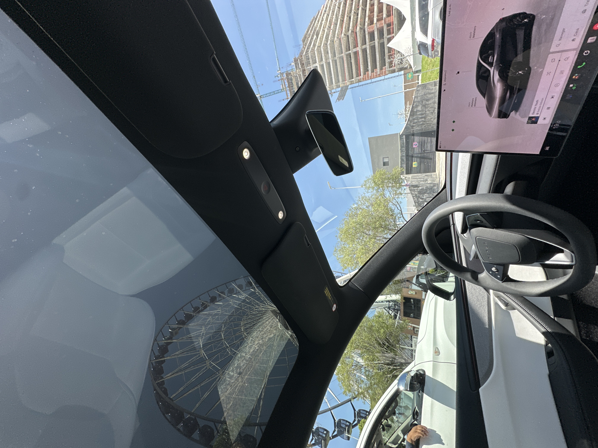 The image captures a view from inside a modern electric vehicle, likely a Tesla, looking towards the expansive glass sunroof. Through the sunroof, a large Ferris wheel is visible against a bright blue sky, suggesting an amusement park or fair setting. In the distance, a tall, unfinished building with exposed rebar and scaffolding indicates ongoing construction, characteristic of a developing urban landscape. The vehicle's interior features a minimalist dashboard with a large central touchscreen displaying the car's interface and a dark steering wheel. To the left, another white vehicle is partially visible, with a person's arm resting on its window. The overall scene suggests a sunny day in an urban environment. This location can be identified as Puebla, Mexico, based on the presence of the unfinished structure that resembles the construction site of the Oceanic Tower project.