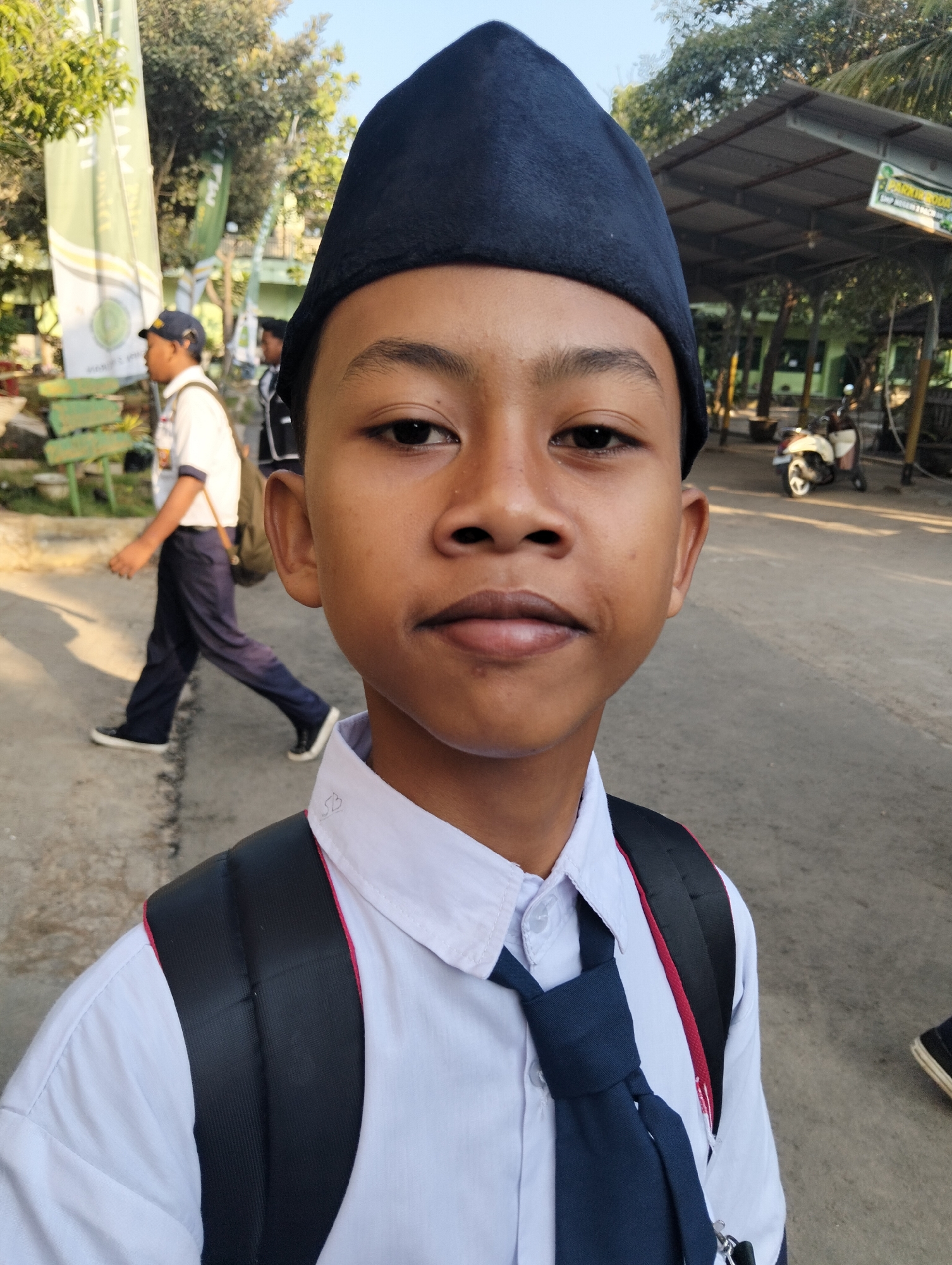 In an outdoor setting, likely a school or public area in Paciran, Indonesia, a young boy in a white shirt, dark blue tie, and a dark, conical cap is the primary subject, captured in a close-up selfie. He wears a black backpack with red trim. In the background, to the left, another person in a school uniform, consisting of a white shirt and dark pants, walks away from the camera, also carrying a backpack. Behind them, trees and banners are visible. To the right, a covered structure with a corrugated roof and a parked scooter suggest a functional, possibly institutional, environment. The ground is paved, and sunlight casts shadows, indicating it is daytime. The overall atmosphere is casual and everyday.