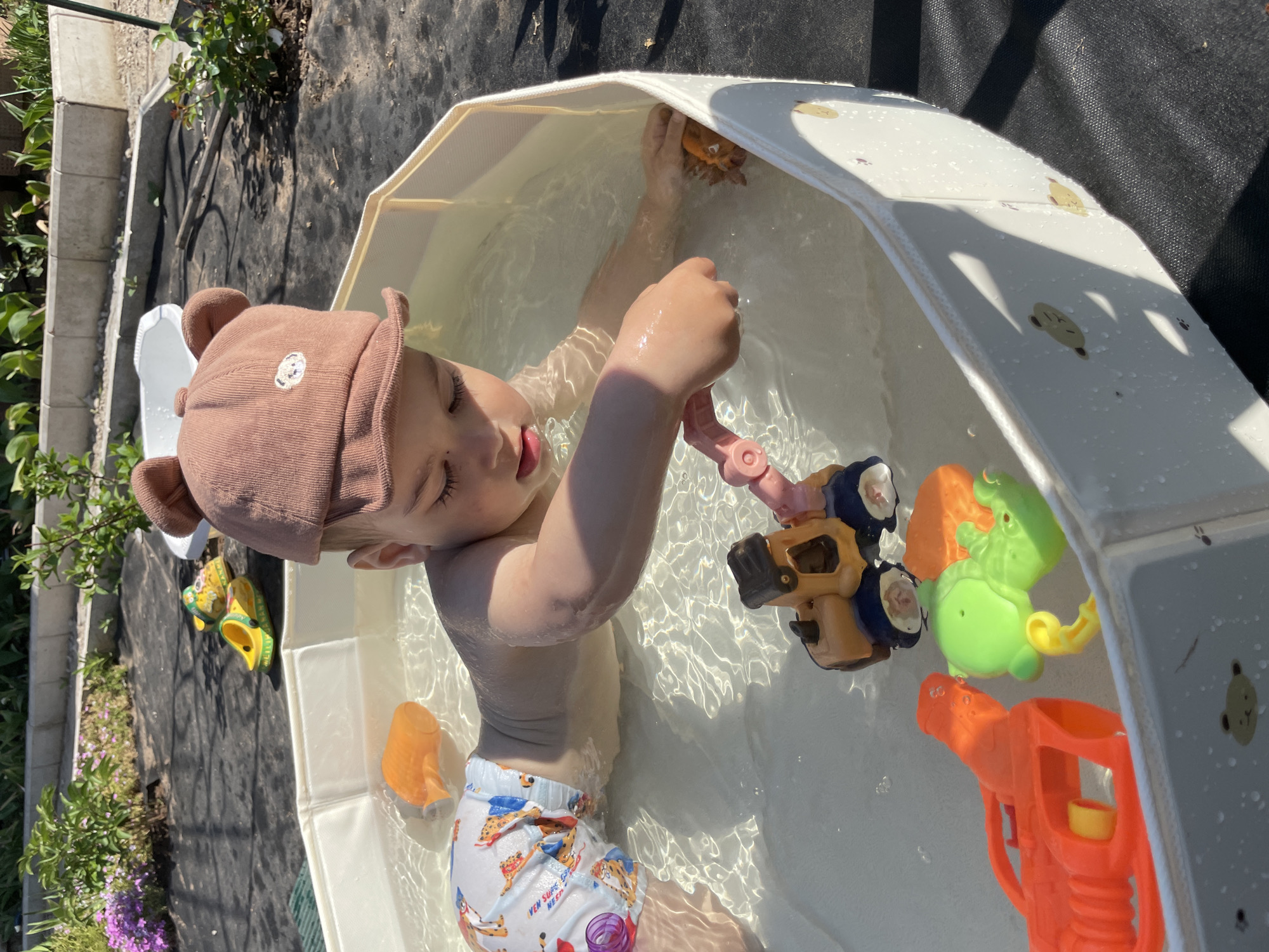 A toddler wearing a brown hat with bear ears and patterned swim trunks is playing in a white plastic pool filled with water. The pool is located outdoors on a sunny day. The toddler is surrounded by several toys, including a toy car, a green alligator, a toy crab, an orange water gun, and an orange cup. The surrounding environment includes garden plants, black woven fabric, and a concrete border. It is not possible to confirm the location as Zav'yalovo, Russia.