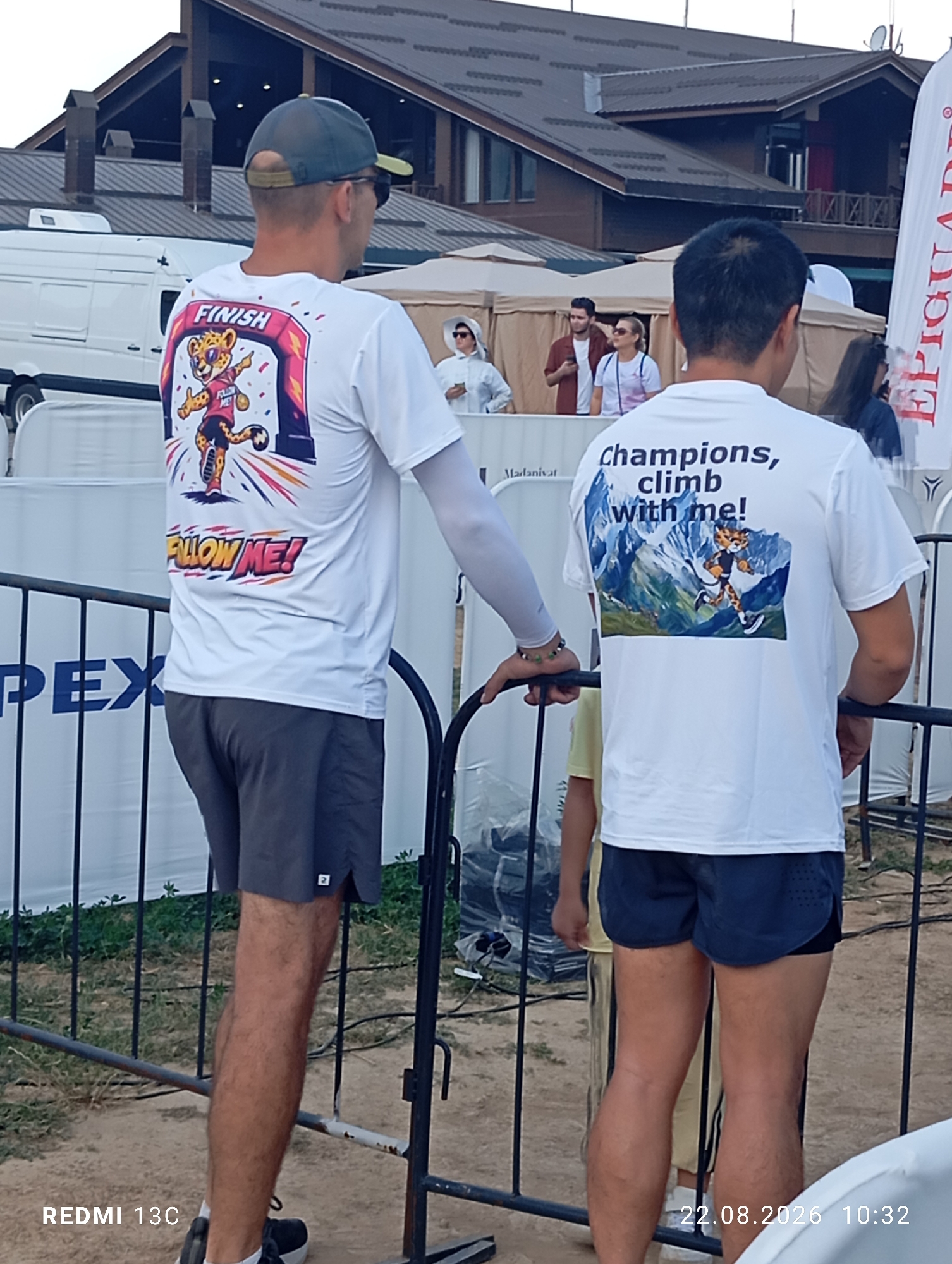 Two men are standing at an outdoor event, likely a race or competition, in Amirsay Ski Resort, Uzbekistan. Both men are wearing white t-shirts with graphics on the back. The man on the left has a graphic of a cheetah crossing a finish line with the text "FINISH FOLLOW ME!". He is wearing a grey baseball cap and sunglasses, and grey shorts. The man on the right has a graphic of a cheetah climbing a mountain with the text "Champions, climb with me!". He is wearing dark shorts. They are standing behind a metal fence. In the background, there are tents and a building structure. The overall scene suggests a sporting event in a mountainous resort setting.