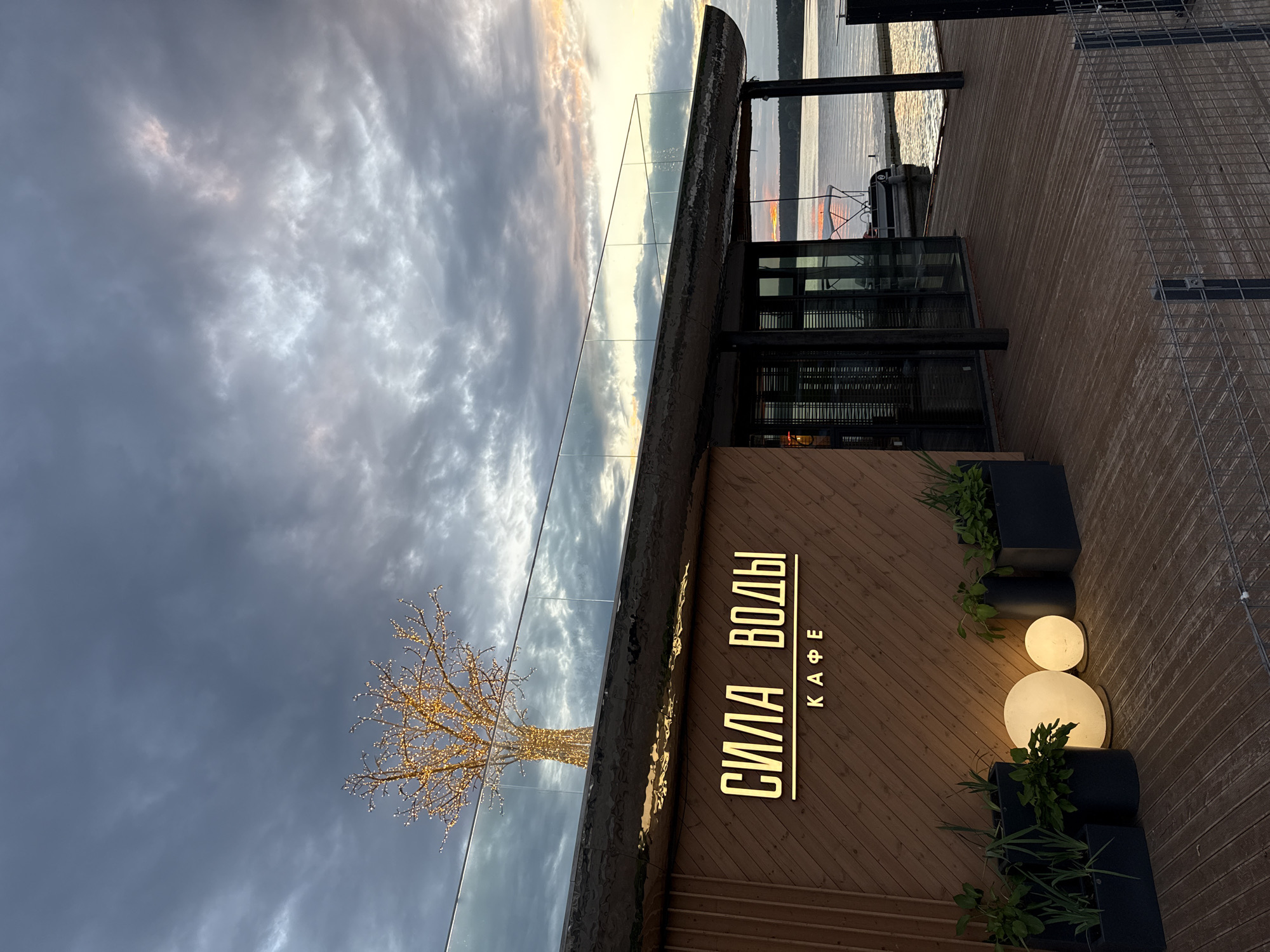 The image depicts a waterfront cafe named "СИЛА ВОДЫ КАФЕ" (Sila Vody Cafe) in Votkinsk, Russia. The cafe has a wooden facade with illuminated signage. In front of the cafe, several planters with greenery are arranged, along with two spherical light fixtures. A modern glass railing runs along the rooftop, where a decorative tree adorned with lights is visible against a cloudy sky. The cafe overlooks a body of water, likely a lake or river, with a boat docked nearby. The scene is captured at dusk, with the sky displaying soft, diffused light from the setting sun. The overall atmosphere is serene and contemporary.