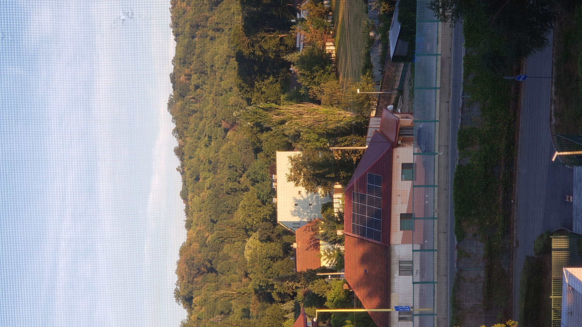 The image captures a suburban scene in Prešov, Slovakia, likely taken from an elevated position, possibly a window with a protective mesh. The foreground features the rooftop of a building with visible solar panels. Adjacent to this is another building with a red tiled roof, and further back, a larger, modern white building. The middle ground reveals a road with a green noise barrier along its side, covered in dense green ivy. Beyond the road, there are more residential buildings and a treed area that slopes upwards. The background is dominated by a dense, rolling hillside covered in lush green trees, suggesting a natural or park-like setting surrounding the developed area. The sky is a clear blue with scattered white clouds, and the lighting indicates it is daytime, possibly late afternoon given the angle of the sunlight casting shadows. The overall impression is one of a residential area bordering a natural landscape.