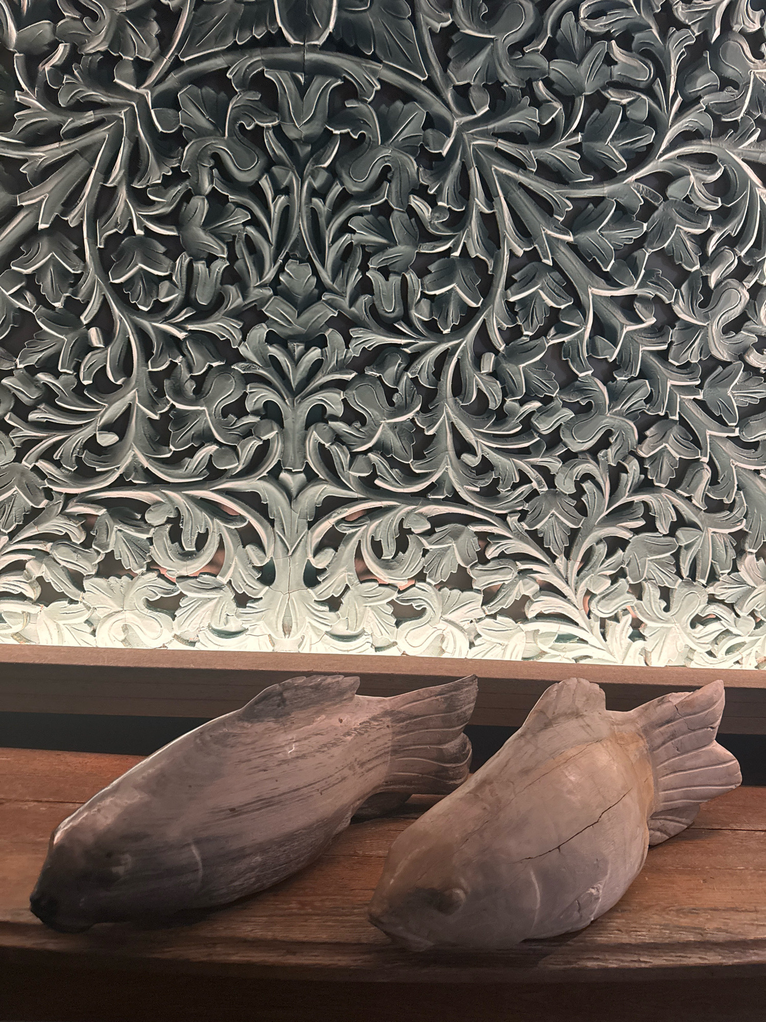 In Agios Tychona Community, Cyprus, two carved wooden fish sculptures rest on a wooden surface. Behind them, a large, intricately carved decorative panel displays a repeating floral and vine pattern in a muted gray color. The panel appears to be backlit, with a soft glow emanating from behind it, highlighting the detailed relief work. The overall scene suggests an indoor setting, possibly a home or a shop, with a focus on decorative elements.