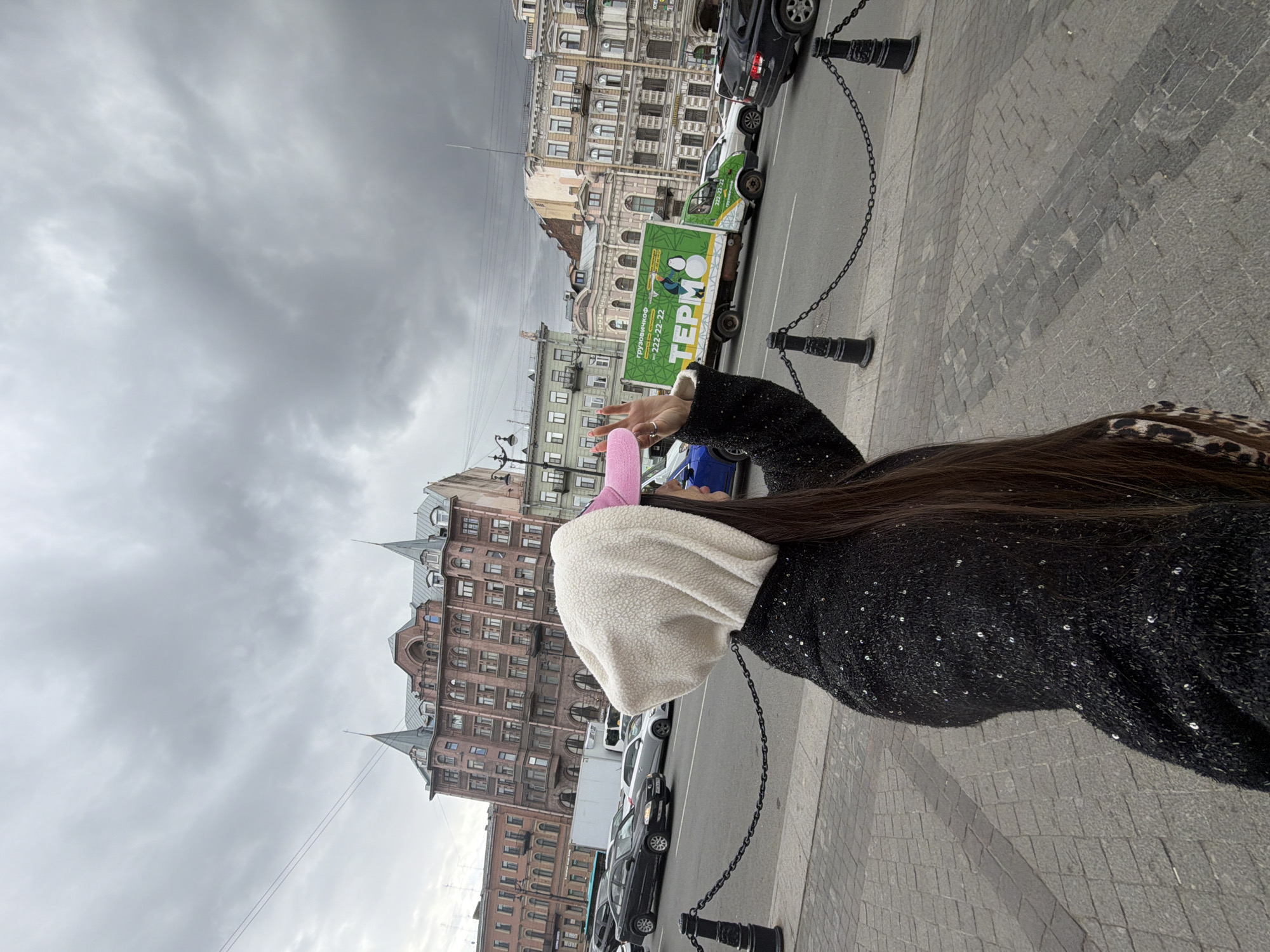 The image depicts an overcast daytime scene on a city street, likely in Saint Petersburg, Russia. A person, seen from behind or the side, stands on a cobblestone sidewalk in the foreground on the right. They have long, dark hair and wear a black top or jacket with subtle reflective embellishments. A light-colored, textured hoodie or hat covers their head, with a pink hat or accessory visible underneath. Their right hand is raised, palm facing outwards, in a gesture that might be shielding their eyes or waving.
The sidewalk is separated from the road by a black chain barrier with decorative posts. On the street, several vehicles are present, including a dark SUV and a partially visible blue car. A prominent green truck with a large white cargo box is clearly visible, adorned with Cyrillic text: "ГРУЗОВИЧКОФФ" (Gruzovichkoff), a phone number "222-22-22", and "ТЕРМО" (TERMO), indicating a moving or delivery service, possibly for temperature-controlled goods.
In the background, a row of historic, multi-story buildings with classic European architecture lines the street. These structures feature numerous windows, ornate detailing, and some possess distinctive pointed spires or roofs, characteristic of older cityscapes. The sky is entirely covered with heavy, grey clouds, creating a muted and subdued atmosphere consistent with overcast weather.