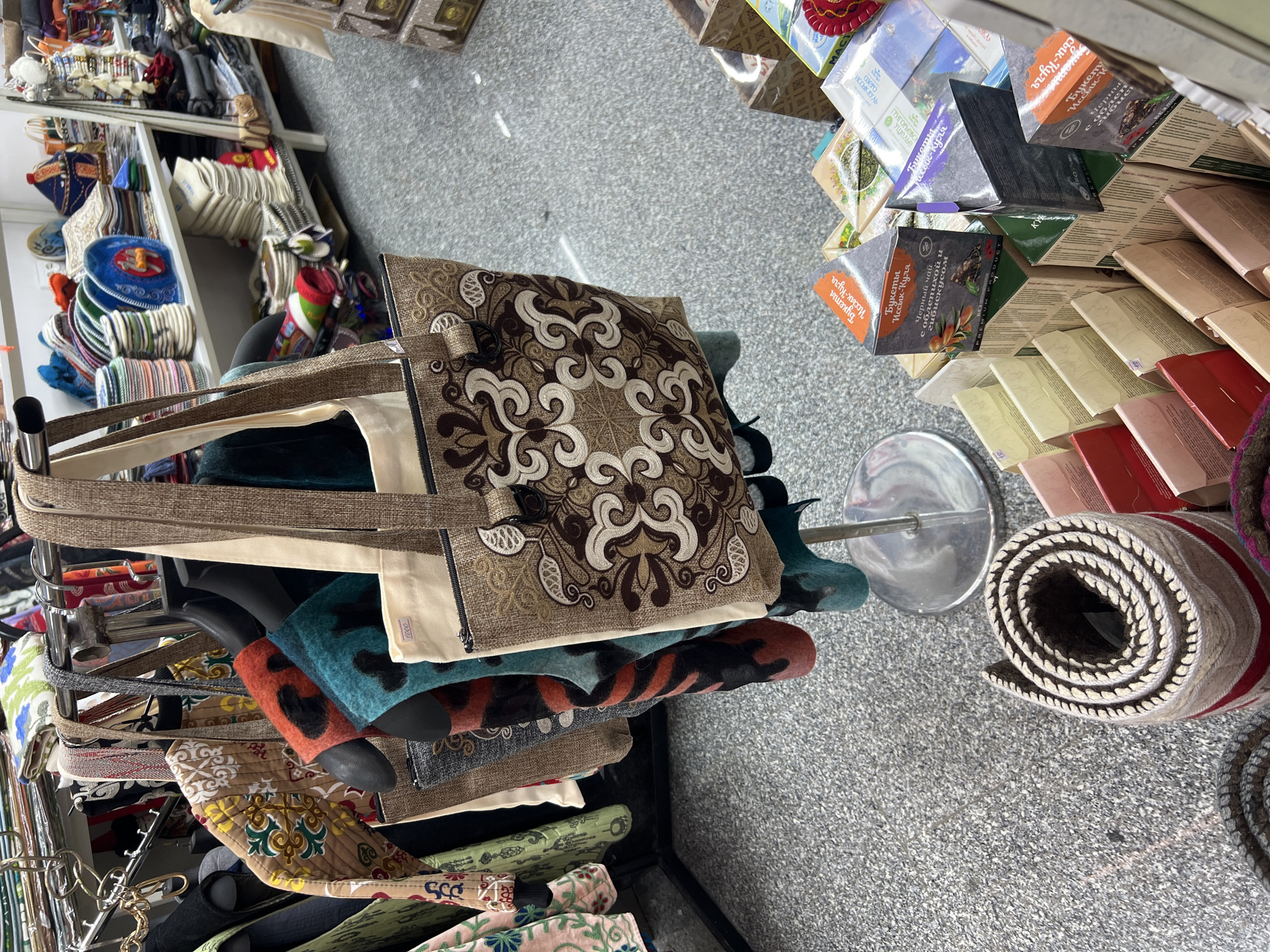 The image depicts a retail display of various textile goods and packaged teas. In the foreground, a large tote bag with an embroidered pattern hangs from a rack. To the right, stacks of tea boxes are arranged in a tiered display. In the background, shelves are filled with folded textiles, hats, and other merchandise. The floor is carpeted with a speckled grey material. A rolling rug is visible in the lower left corner. The scene appears to be an indoor market or shop. The city of Bishkek, Kyrgyzstan, is mentioned as the location.