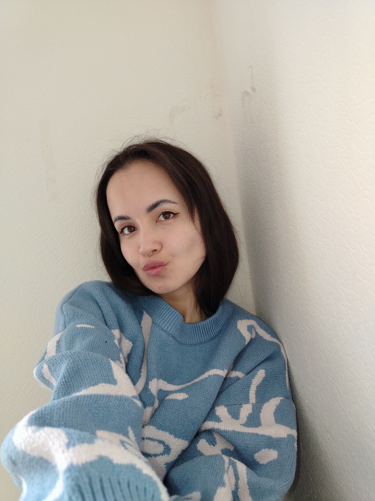 A medium close-up portrait shows a young woman posing indoors in Noviy, Russia. The woman, likely in her twenties, has dark brown, medium-length hair parted on her left side. She is looking directly at the camera with a playful expression, her lips pursed into a 'duck face' or kissing gesture. Her brown eyes are open, and she has dark, well-defined eyebrows with visible eyeliner. She wears two nose piercings: a small golden hoop in her left nostril and a golden stud in her right nostril.
She is dressed in a light blue knitted sweater featuring an abstract white pattern. The sweater appears to be loose-fitting, with visible ribbing at the neck and cuffs. The background consists of plain, light-colored (off-white or very light beige) walls that have a subtle textured surface, possibly painted plaster or wallpaper. A visible corner on the right side of the frame indicates she is positioned against a wall junction. The lighting is bright and even, suggesting daytime. Some faint smudges or marks are visible on the wall above her head. No visible text is present in the image. The overall mood is casual and lighthearted.