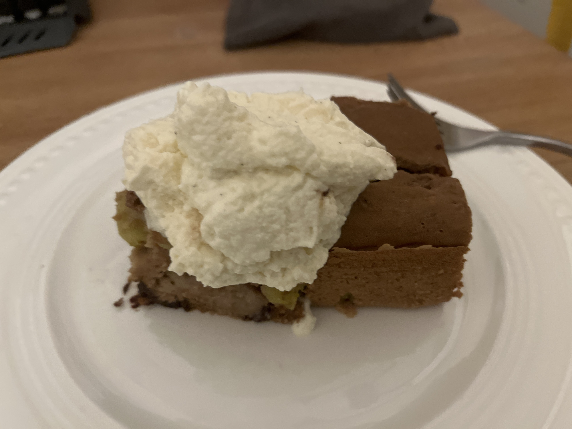 A close-up shot shows a slice of cake topped with whipped cream on a white plate. The cake is brown and appears to have fruit mixed in. A fork is visible to the right of the plate, resting on the surface. The background consists of a wooden table and a dark, blurry object. The scene is indoors, likely in a domestic setting, as indicated by the tableware and table surface. The lighting is warm, suggesting it may be artificial or from a window.