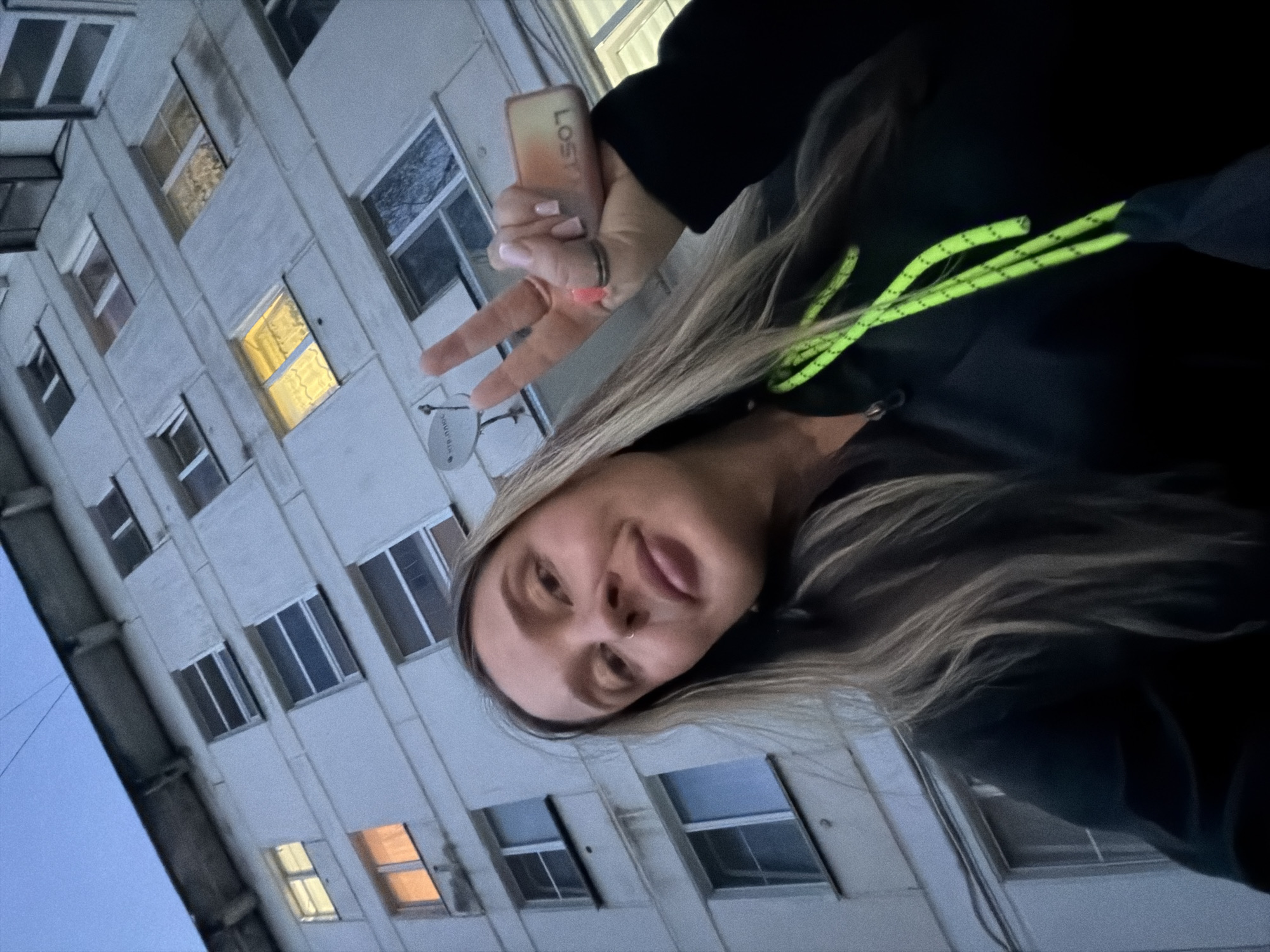 A woman is posing outdoors in what is likely an urban environment. The background features an apartment building with multiple windows, some with lights on. The woman is giving a peace sign. She is wearing a black hoodie with neon green drawstrings. She appears to be holding a disposable vape. The image was taken at dusk. It is highly probable the location is Chaikovsky, Russia, given the building architecture typical of the region.